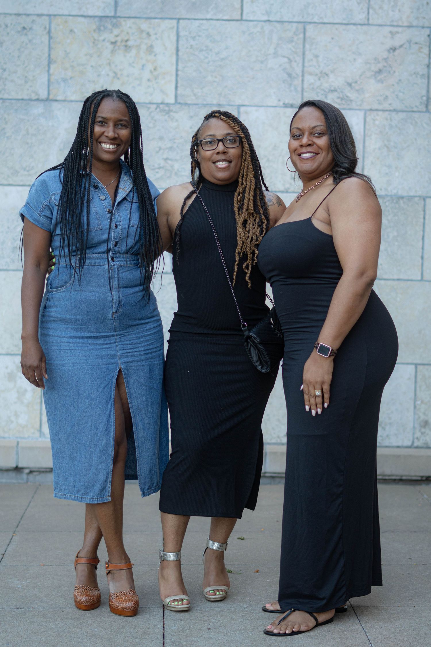 Three women in stylish dresses pose together against a stone wall, wearing blue denim and black outfits.