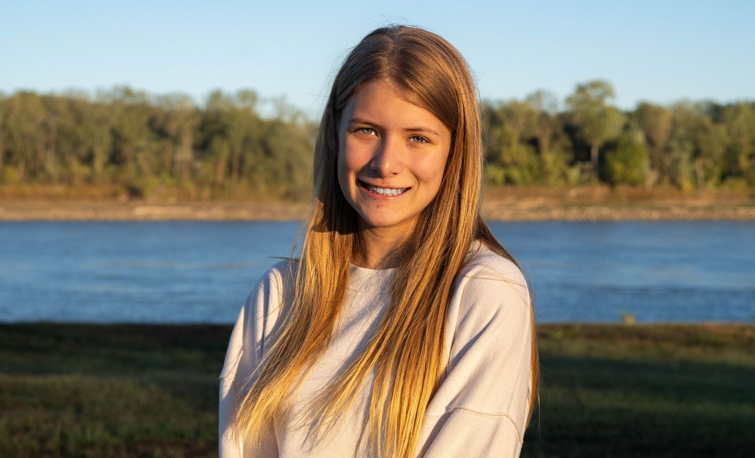 Woman with long blonde hair poses outdoors by a lake at sunset with natural scenery in background.
