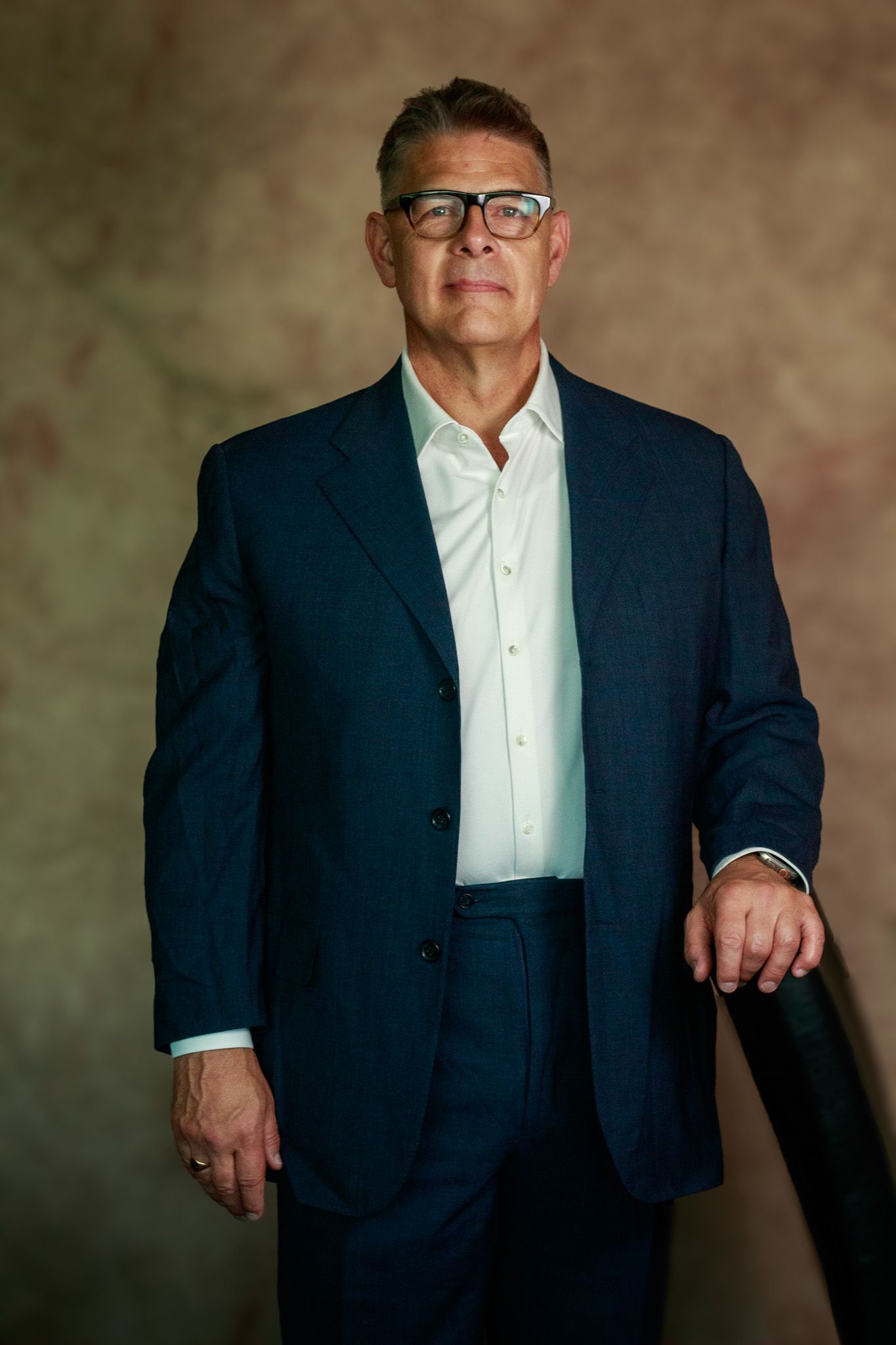 A professional portrait of a businessman wearing glasses and a navy suit against a brown backdrop.