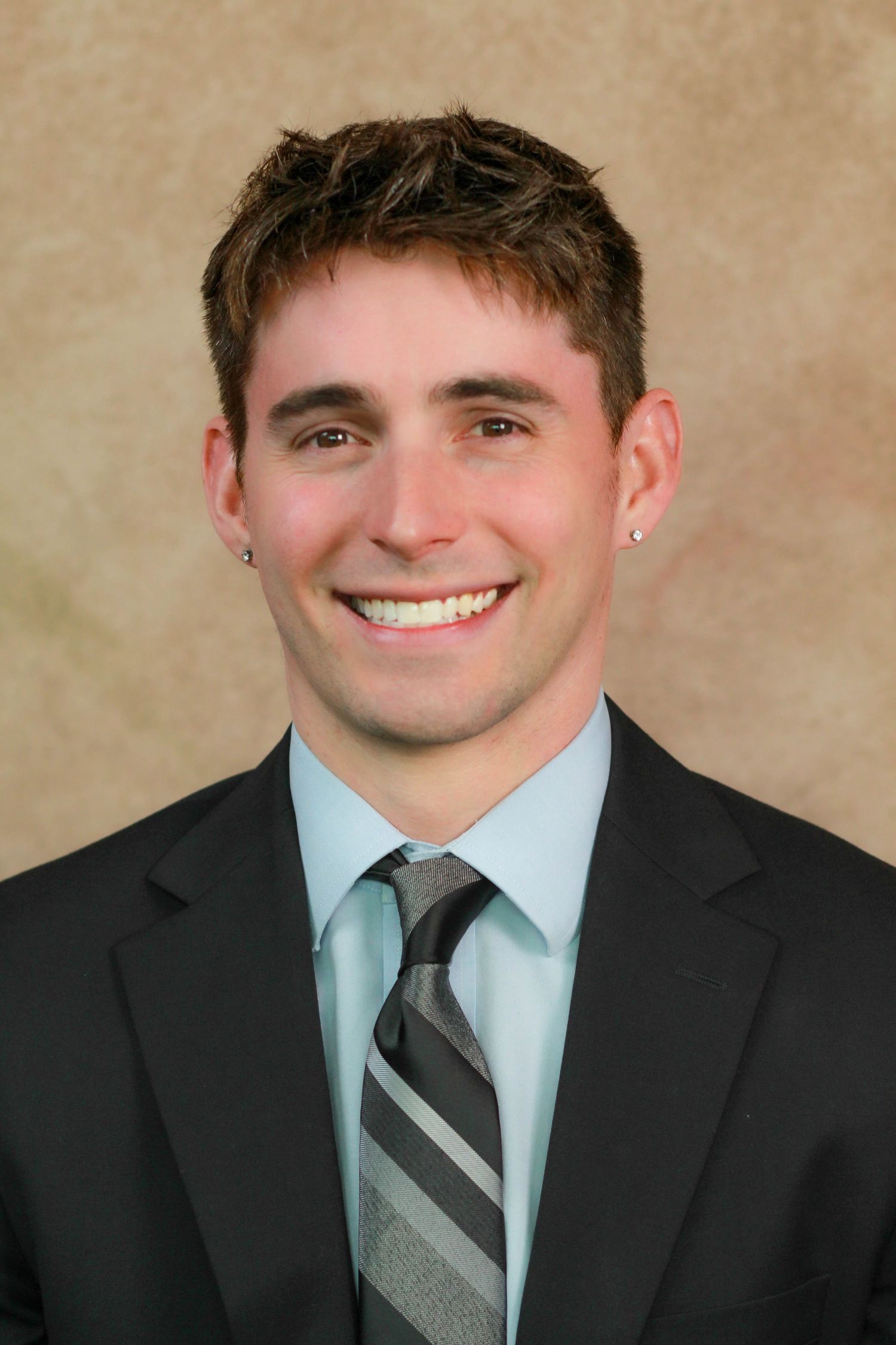 Professional headshot of person in dark suit with striped tie and bright smile.