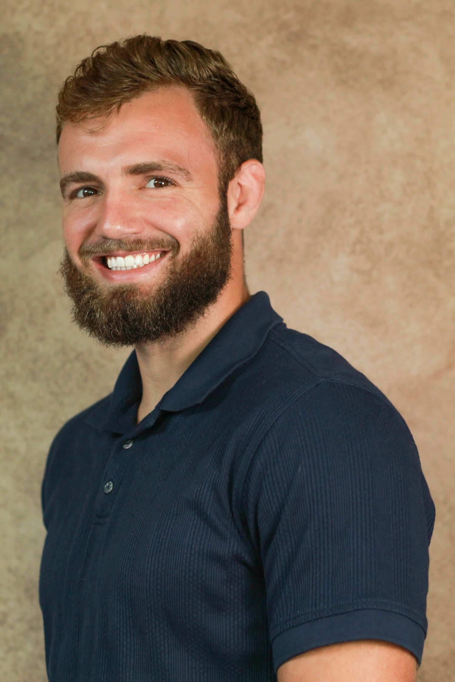 Person with full beard wearing navy polo shirt smiling warmly in professional photo.