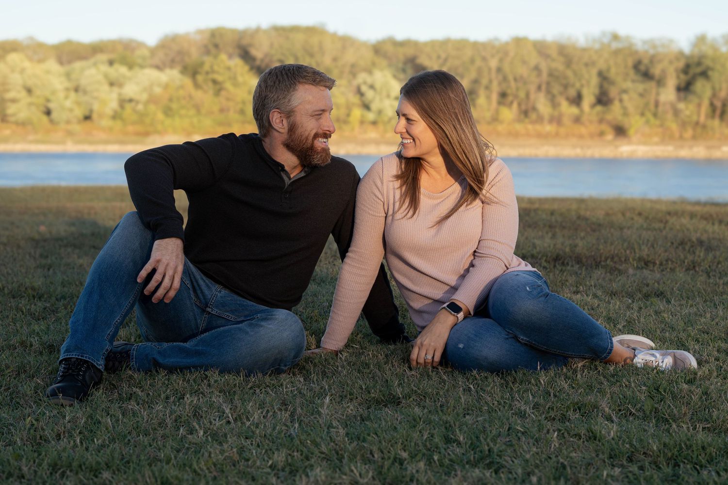 A couple relaxes together on grassy hillside during golden hour sunset in romantic outdoor photoshoot.