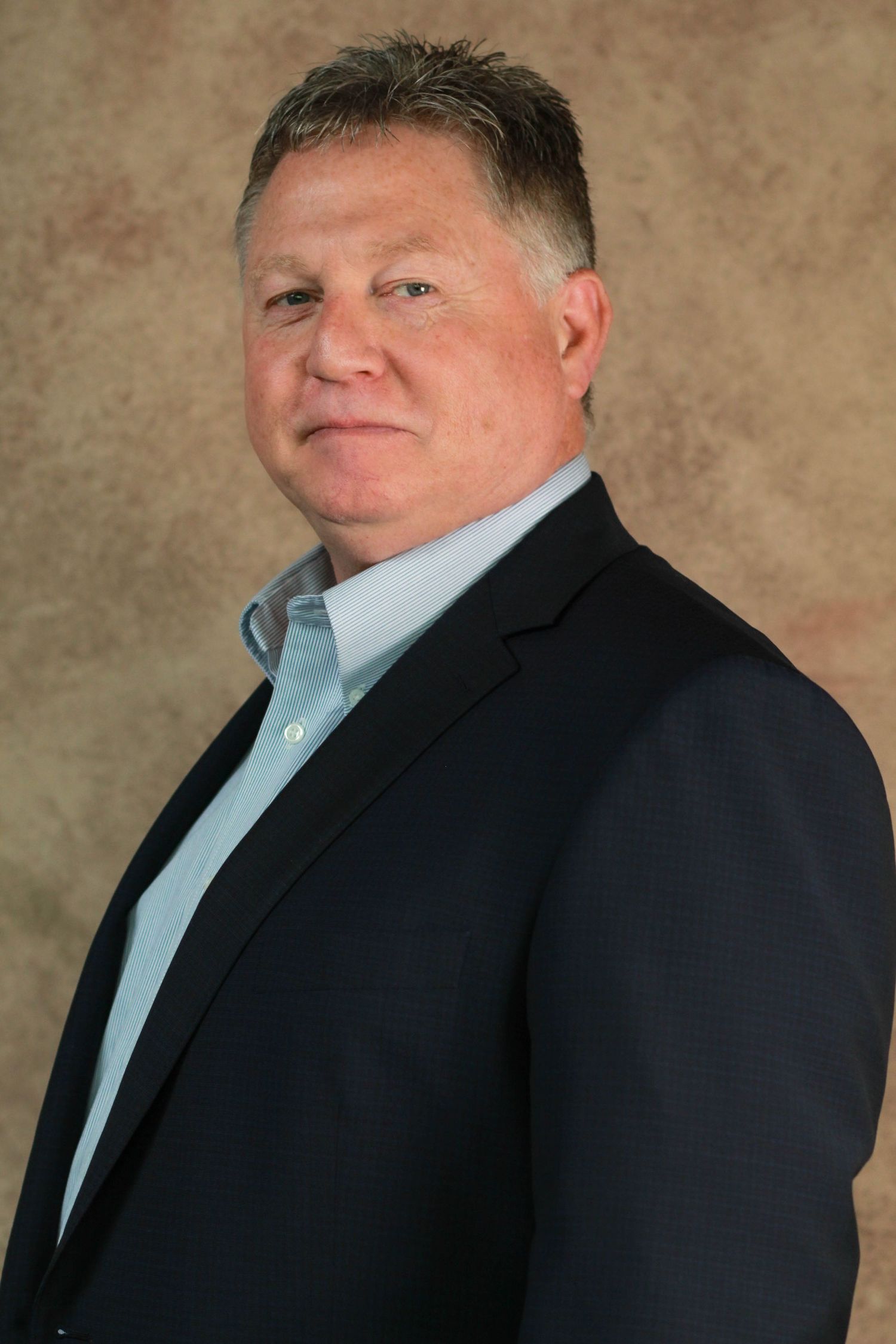 Professional portrait of mature person in dark blazer and light blue shirt against textured backdrop.