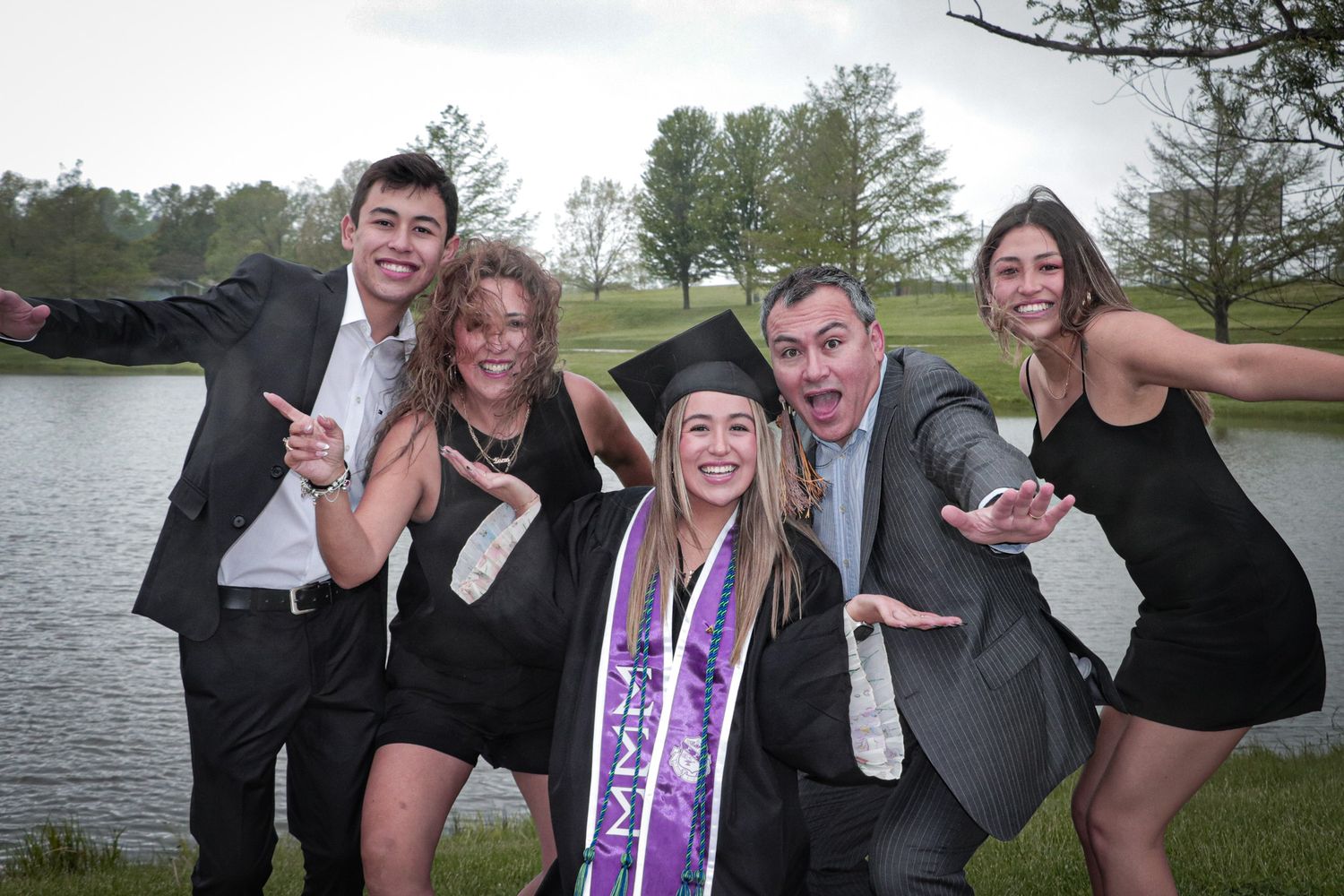 A group celebrates a graduation with playful poses by a lake.