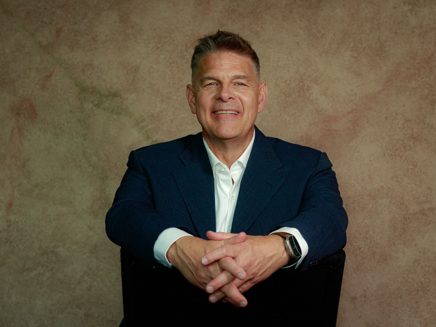 A professional headshot of a businessman in a navy suit against a textured brown background.
