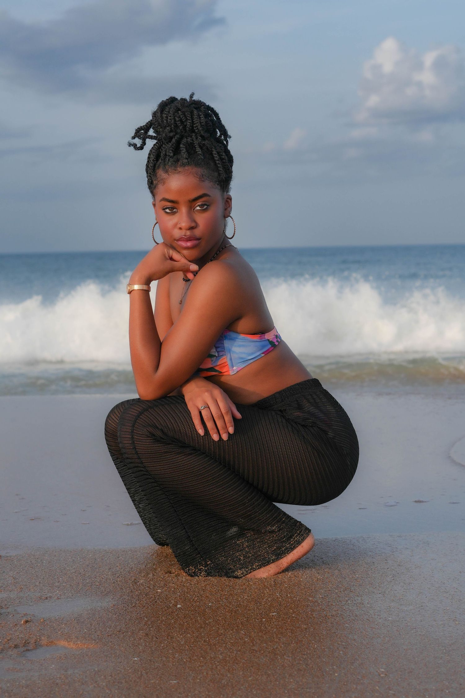 Artistic beach photoshoot with person in black dress sitting on sand with waves crashing behind.