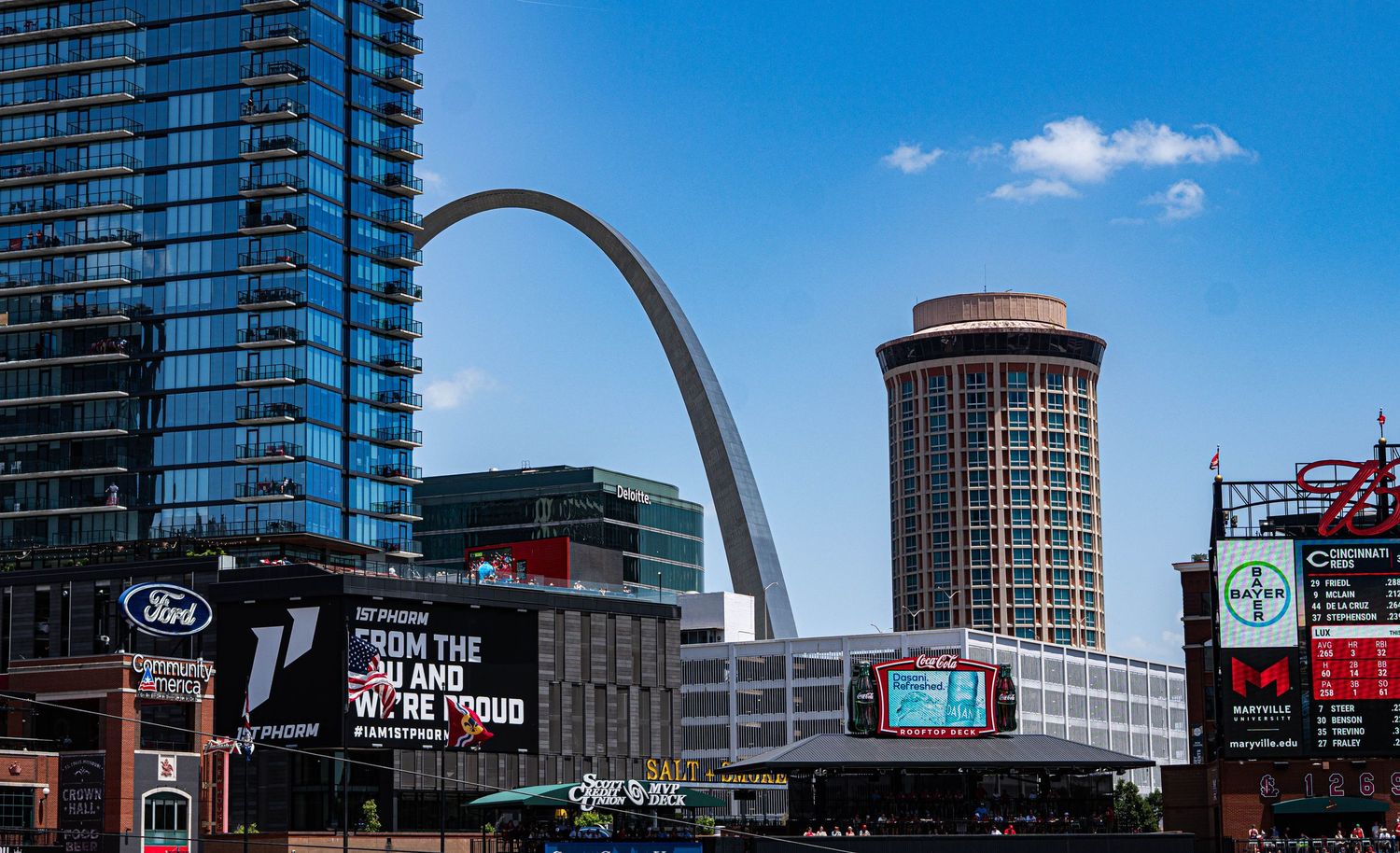 The iconic Gateway Arch dominates the St. Louis skyline with modern buildings and signage below.