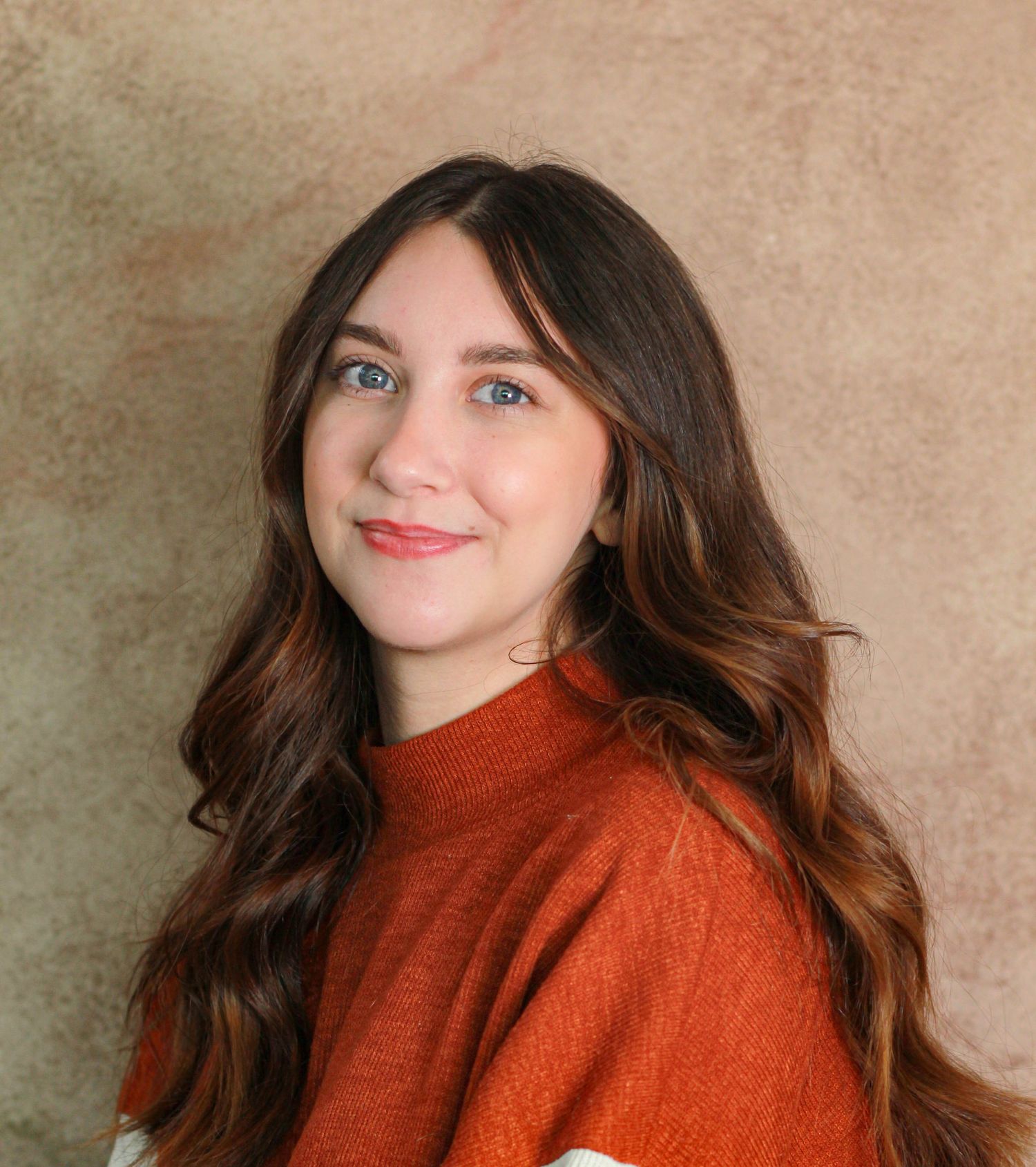 Person with long wavy brown hair wearing rust-colored sweater in professional portrait.