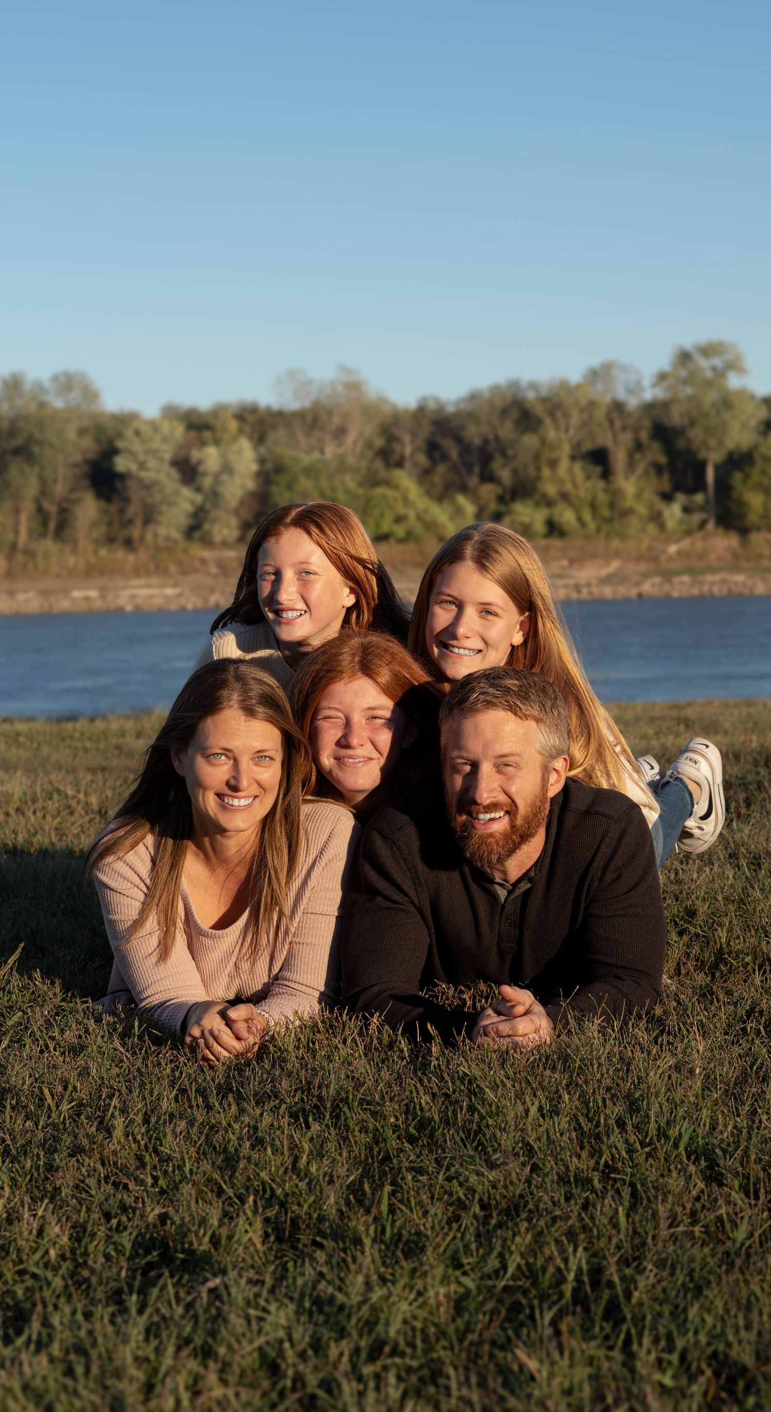 A family group lounges together on grass by a lake on a sunny afternoon with trees in the background.