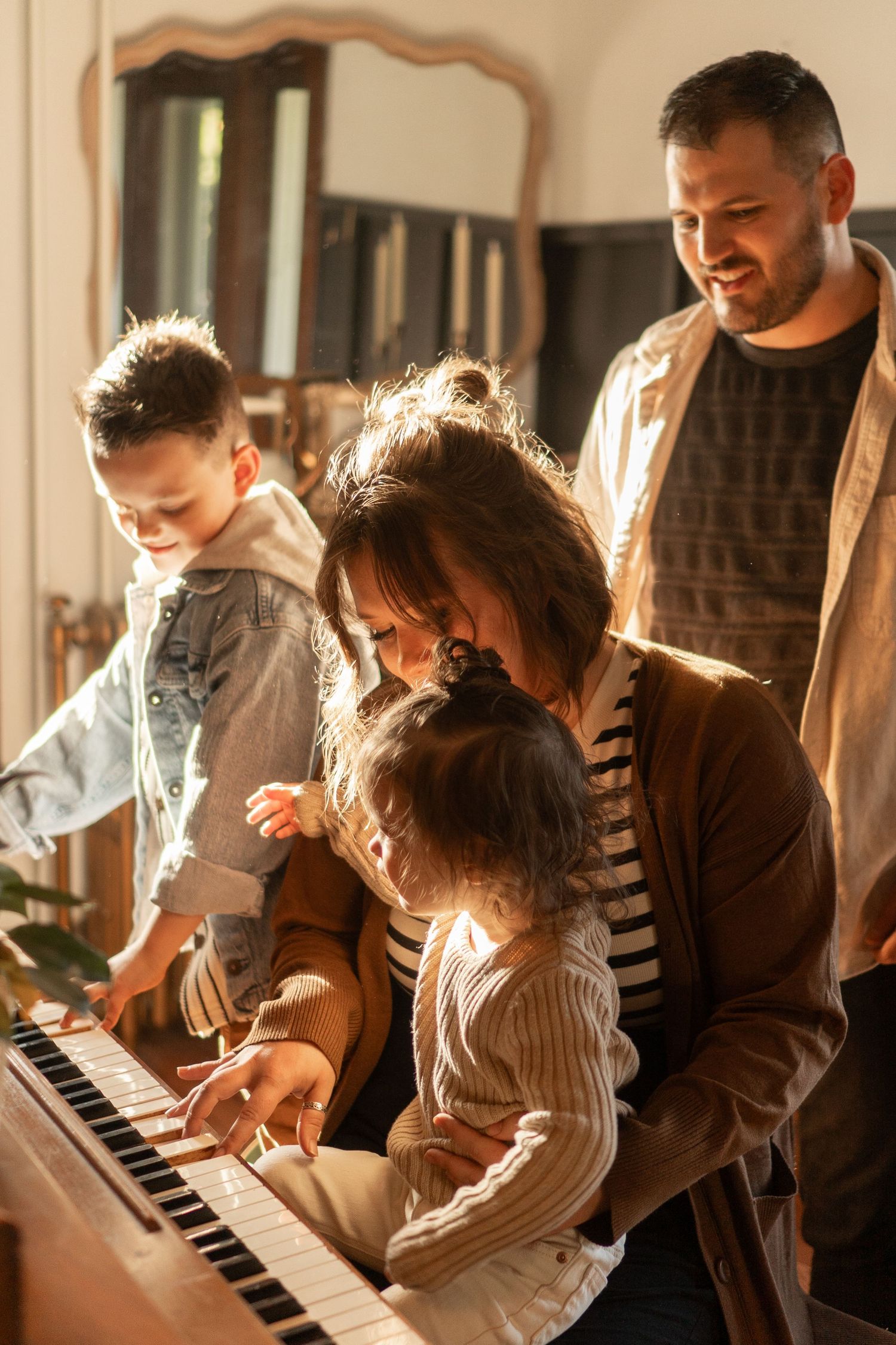 A happy family gathers around a piano, with a mother teaching her young daughter to play while father and son watch.
