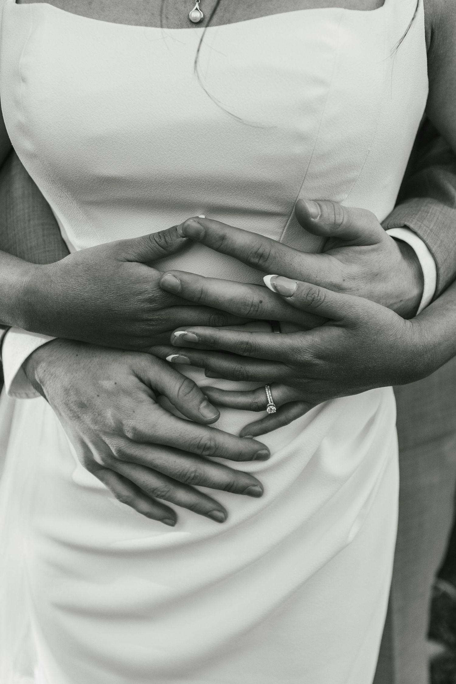 Black and white photo of newlyweds hands clasped together showing wedding rings, bride in white dress.