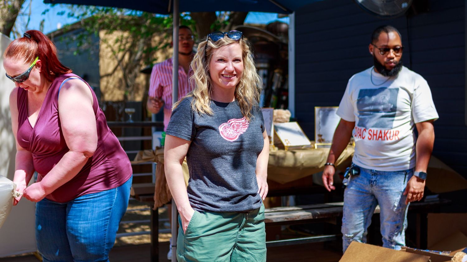 Three people standing outdoors at a community event on a sunny day, smiling and engaged in activities.