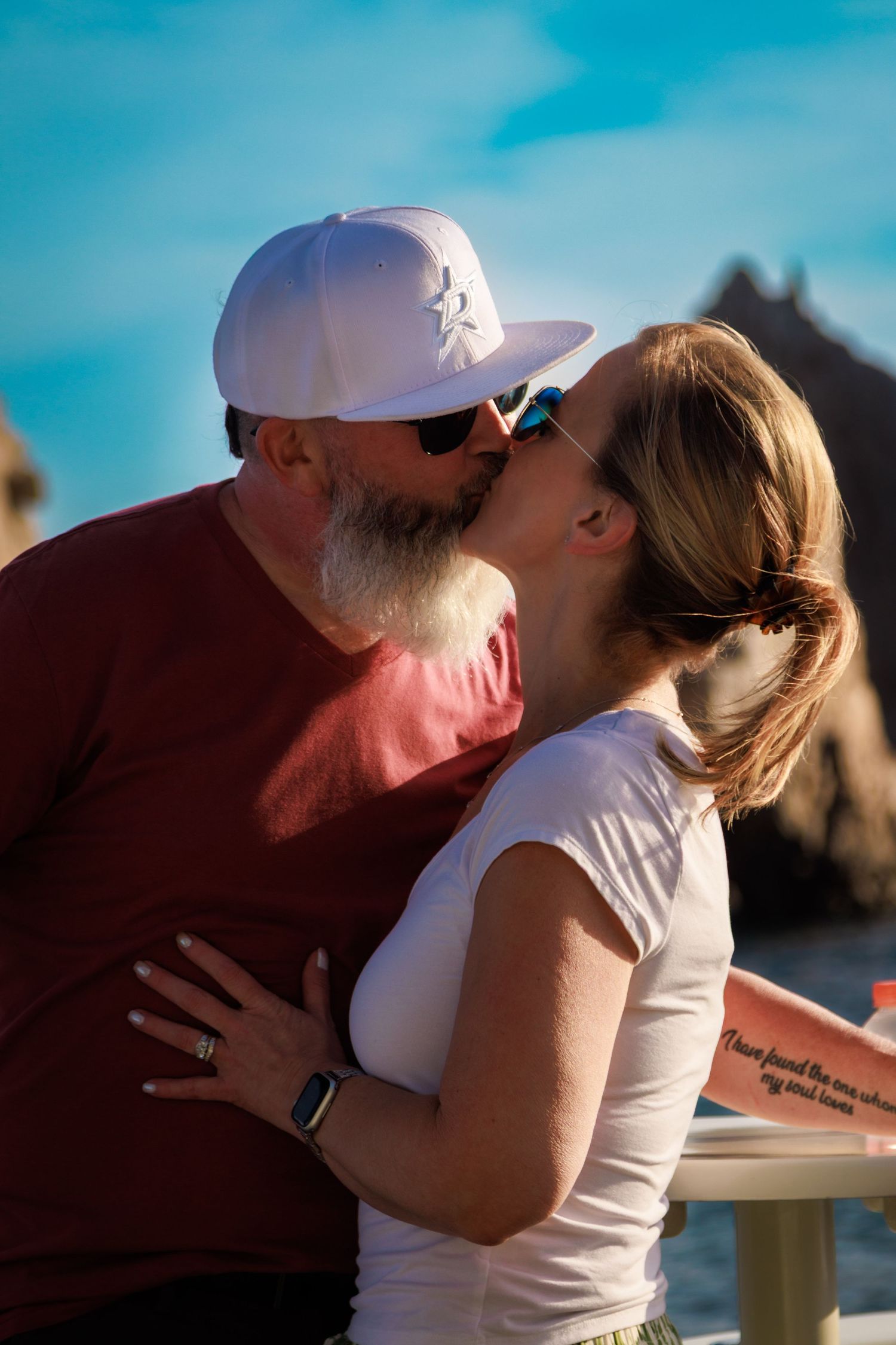 A bearded man in a white cap and sunglasses shares a romantic kiss with a woman outdoors by the ocean.