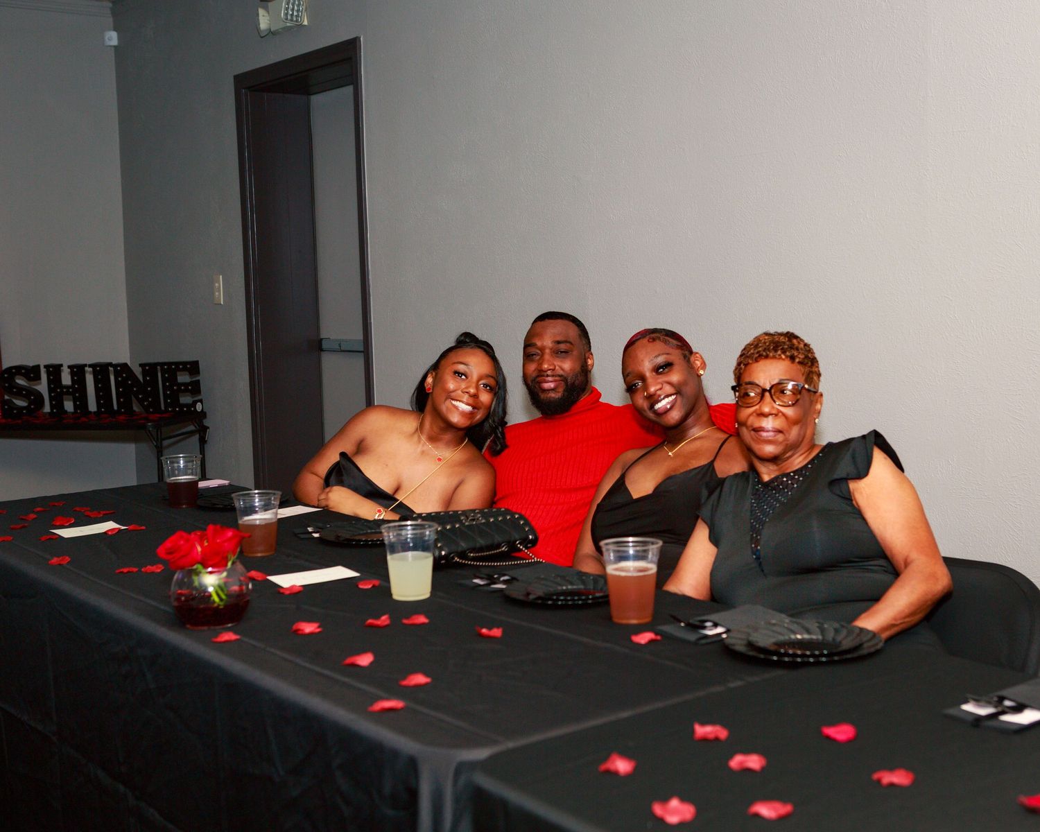 Five people smiling at a decorated black table with red rose petals at a formal event near a SHINE sign.