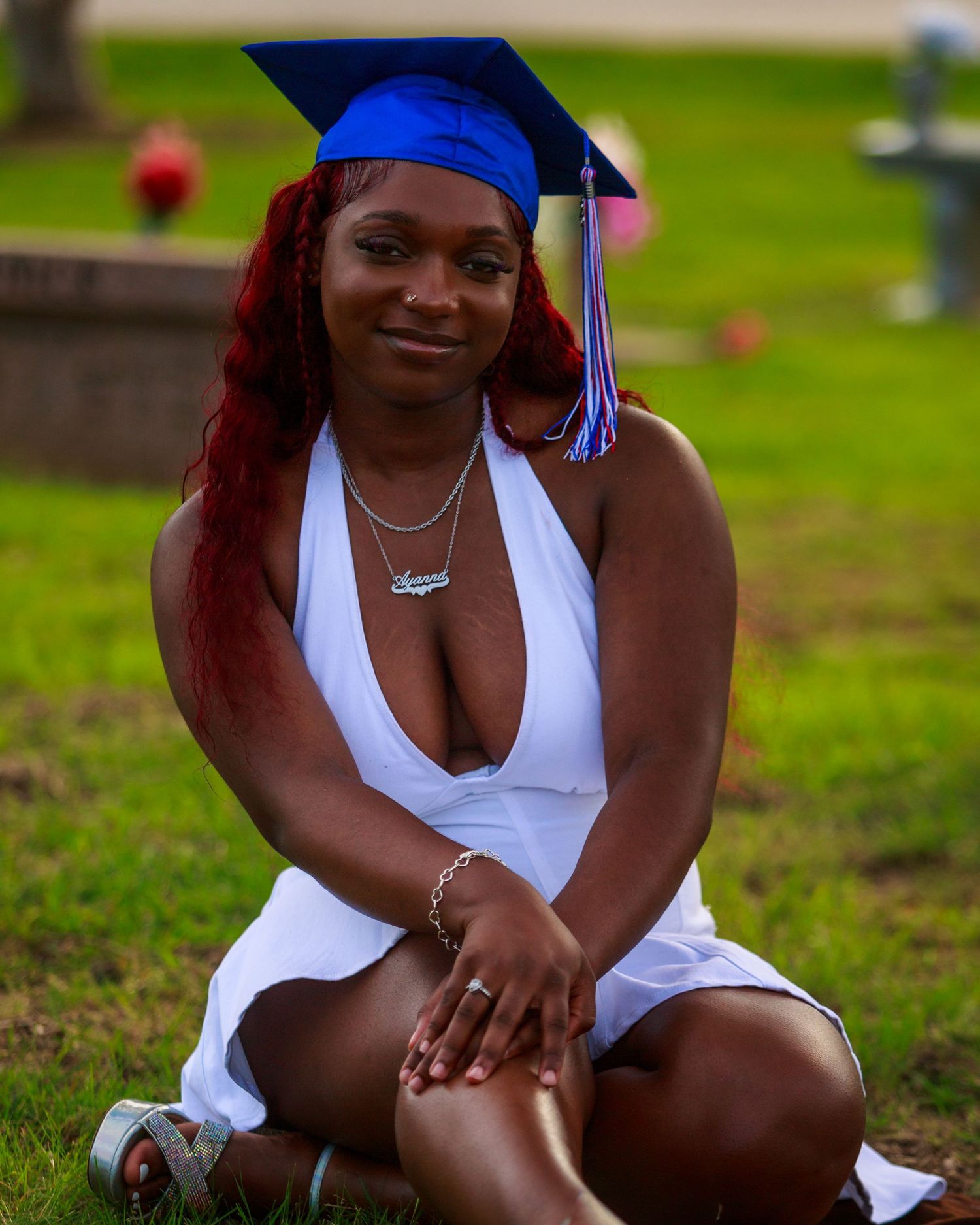Young woman in white dress and blue graduation cap sitting on grass celebrating academic achievement.