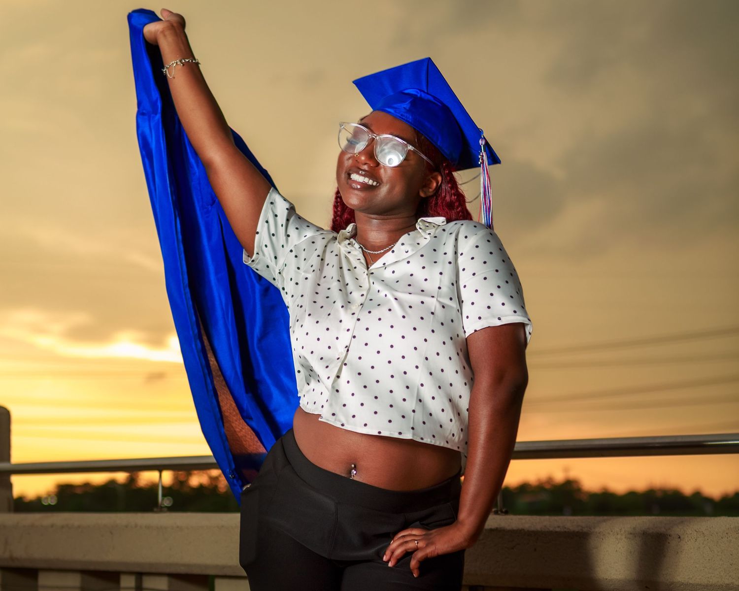 Graduate in blue cap holds gown aloft at sunset, smiling proudly in white polka dot top.