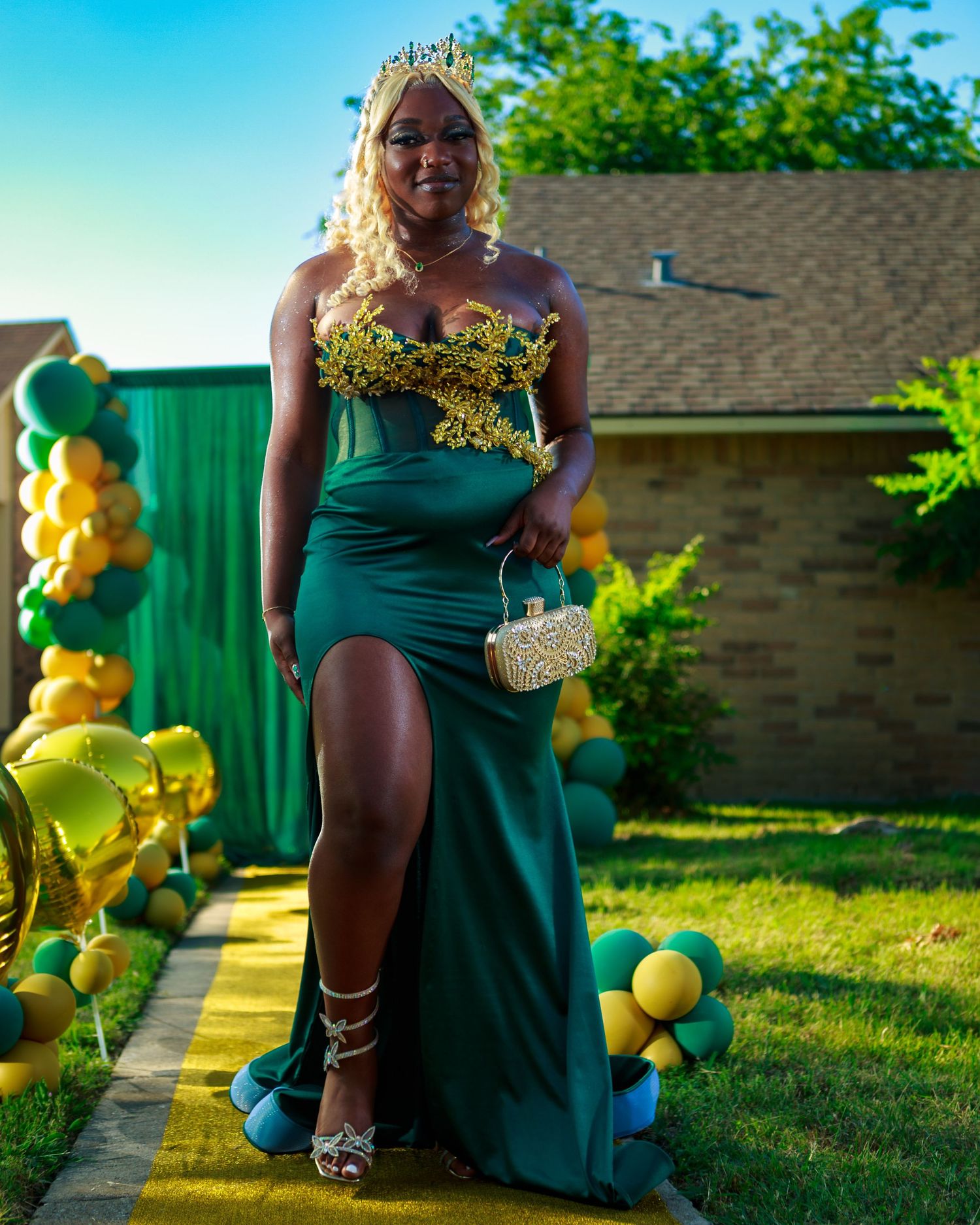 Young woman in elegant green and gold strapless prom gown with crown and clutch, posing outdoors with balloon arch.