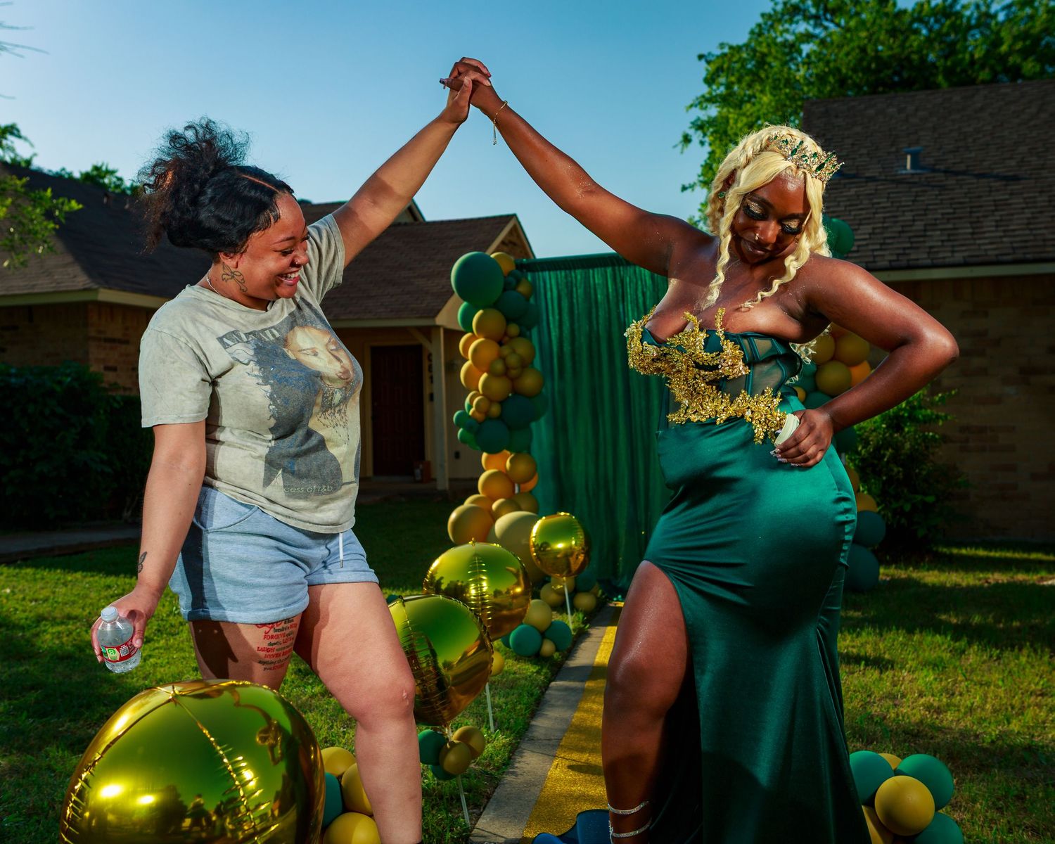 Two women celebrate outdoors, surrounded by green-and-gold balloon decorations, at a festive prom send-off.