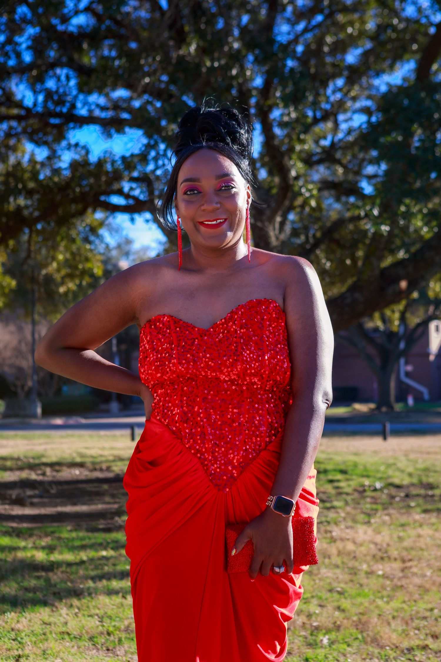 Young woman in red sequin strapless gown posing outdoors under trees.