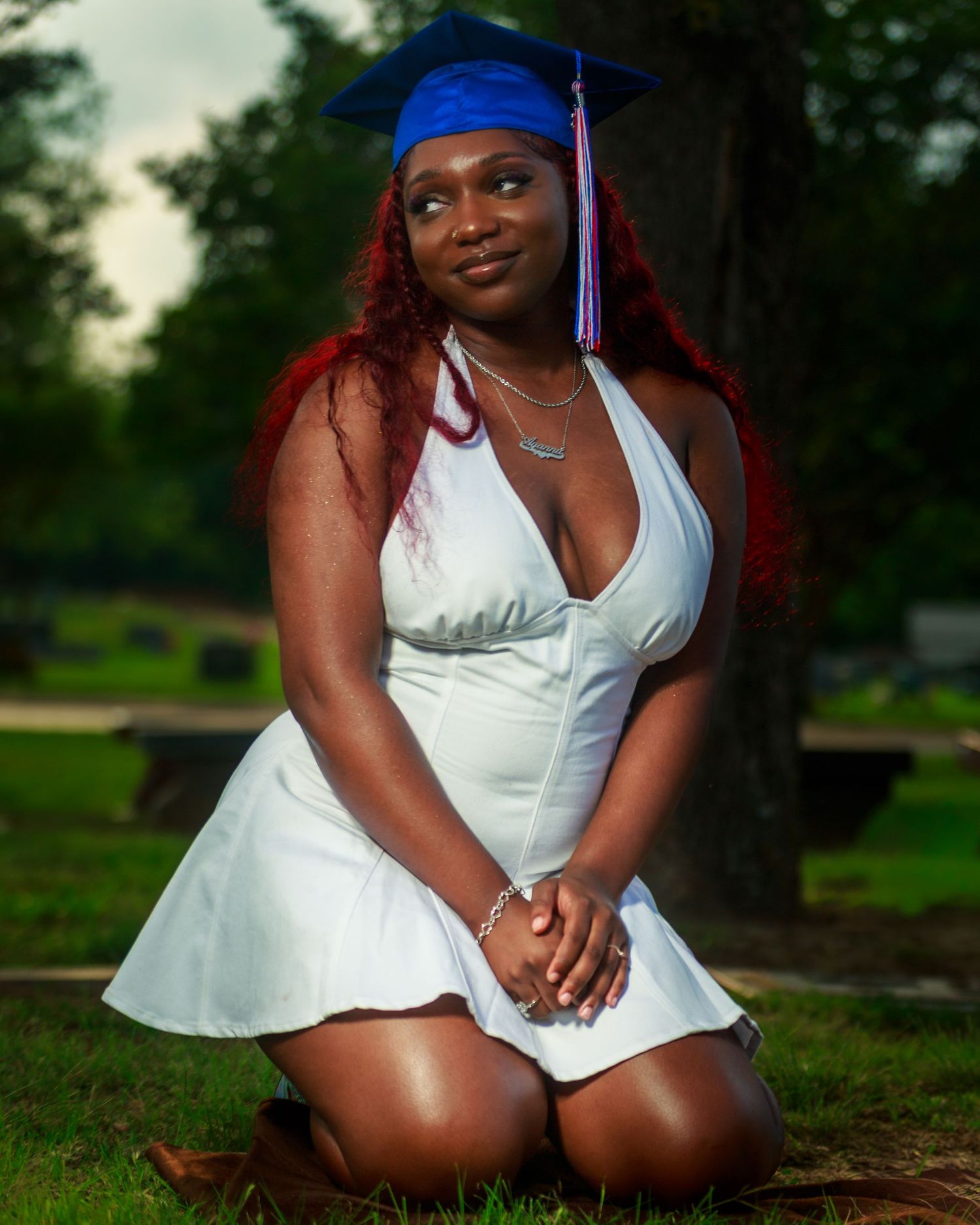 Young woman in white dress and blue graduation cap kneeling on grass for graduation photo.
