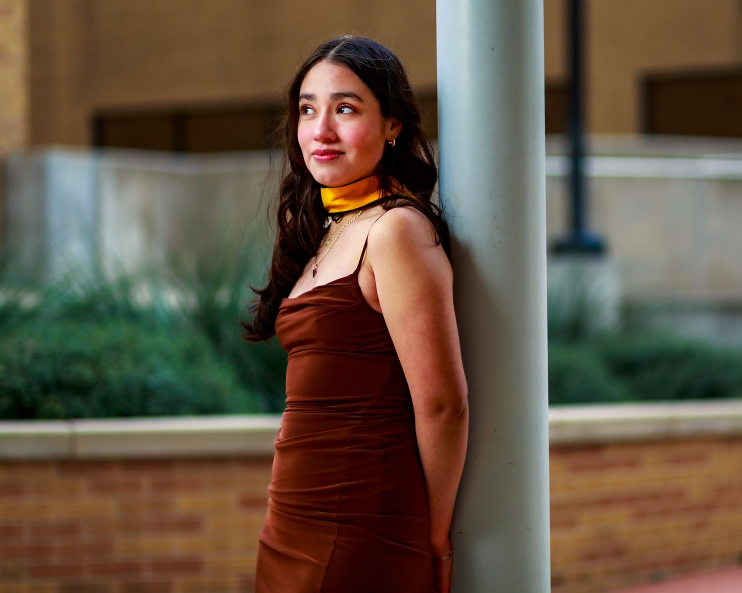 Young woman in brown velvet dress and yellow scarf leans against a pole outdoors in an urban setting.