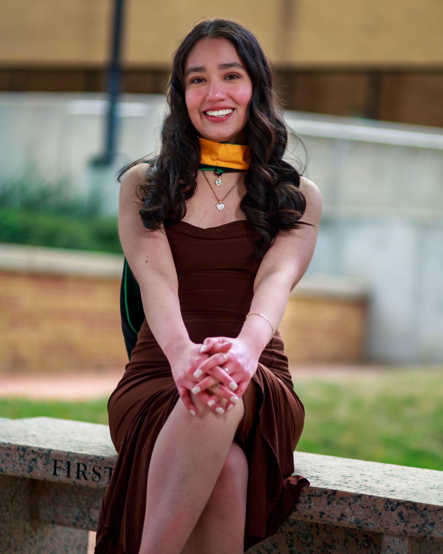 Smiling young woman in brown dress with gold scarf sitting outdoors on stone ledge at graduation.