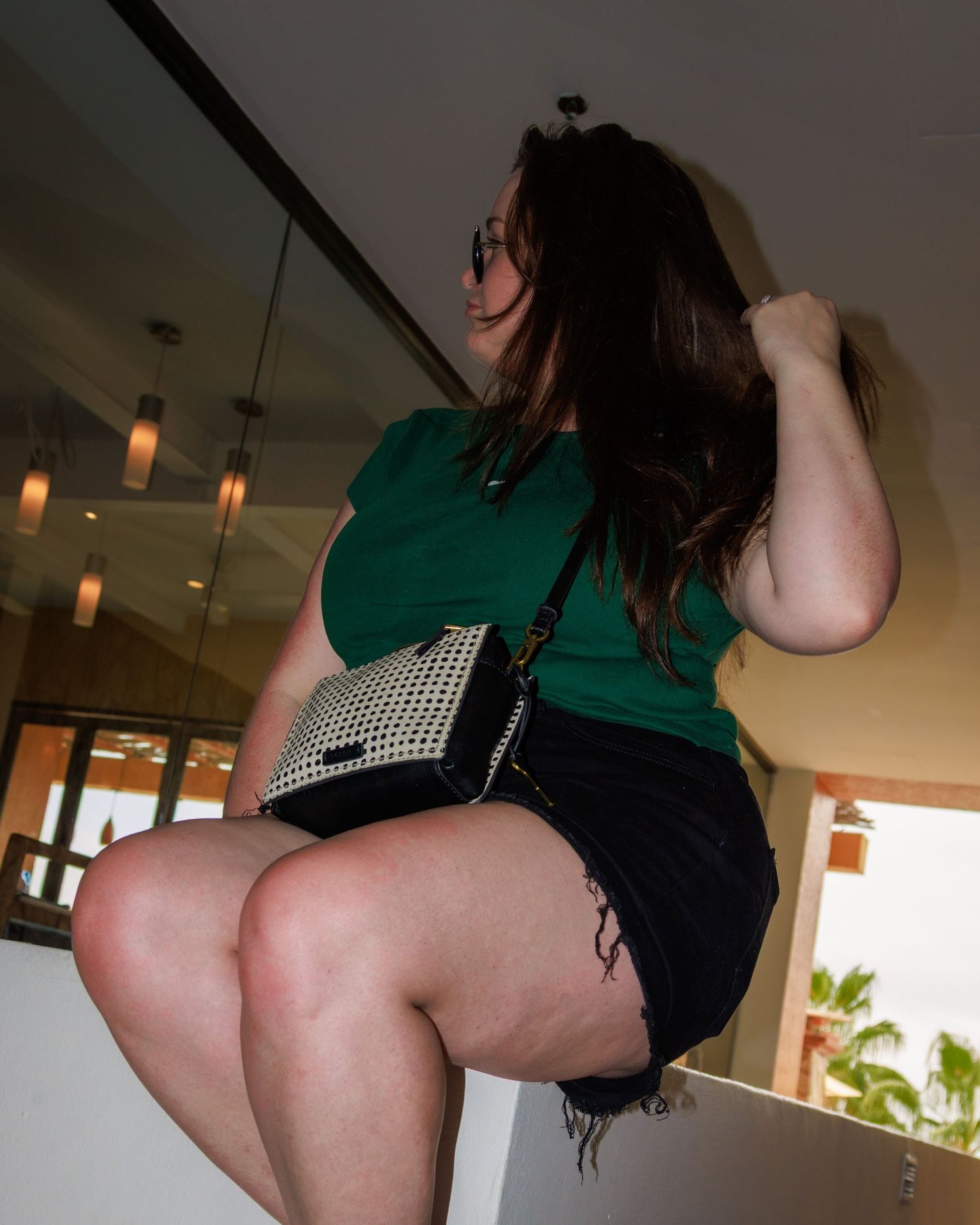 Woman in green top and black mini skirt posing indoors with a woven crossbody bag.