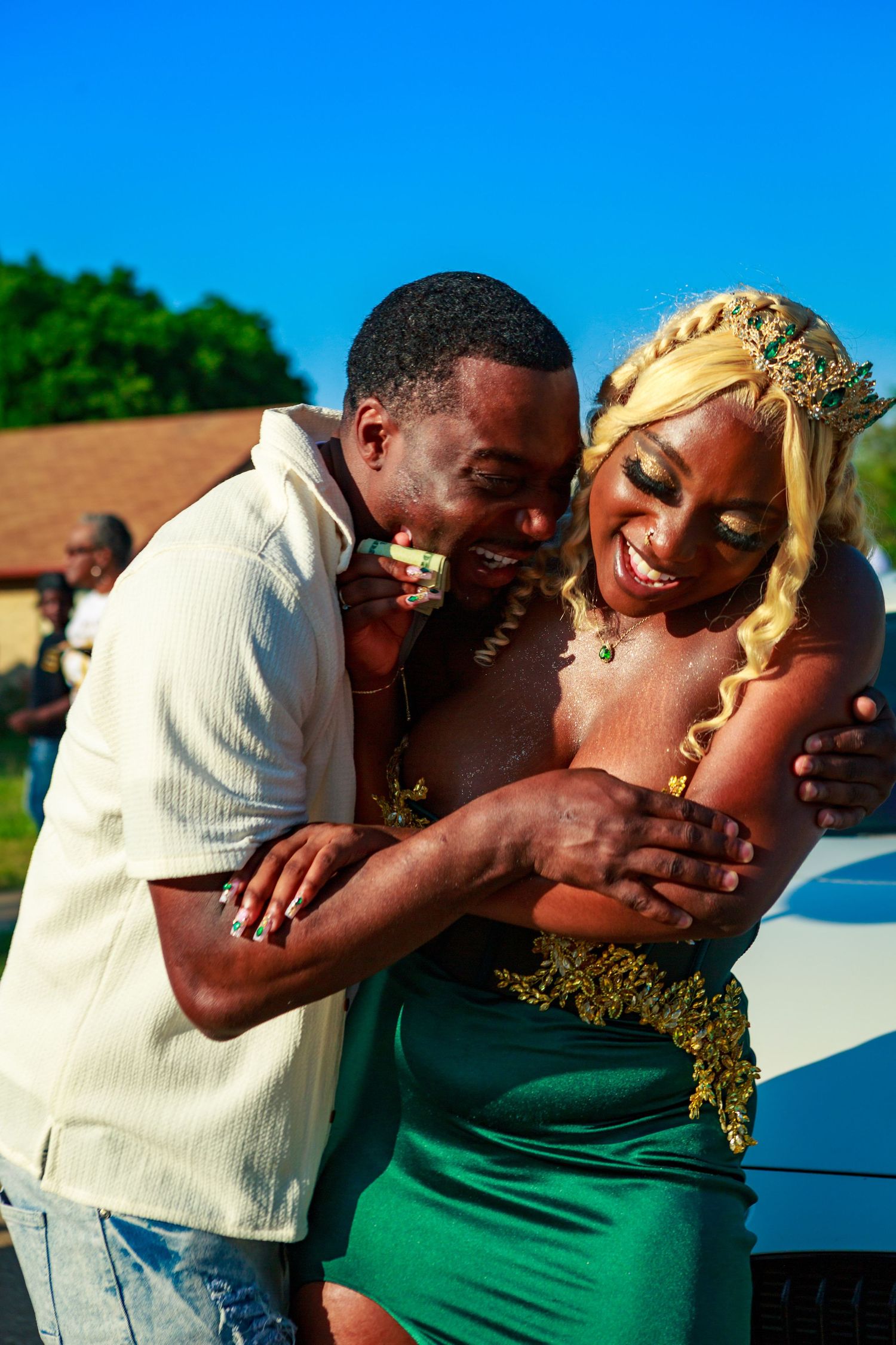 A joyful couple embraces outdoors, woman in green gown with gold crown, man in white shirt, both laughing.