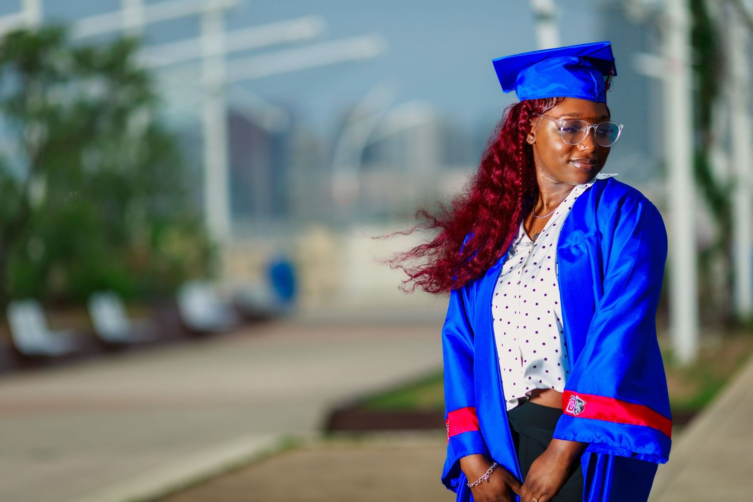 A woman in a blue graduation cap and gown with red hair poses outdoors on a bright sunny day.