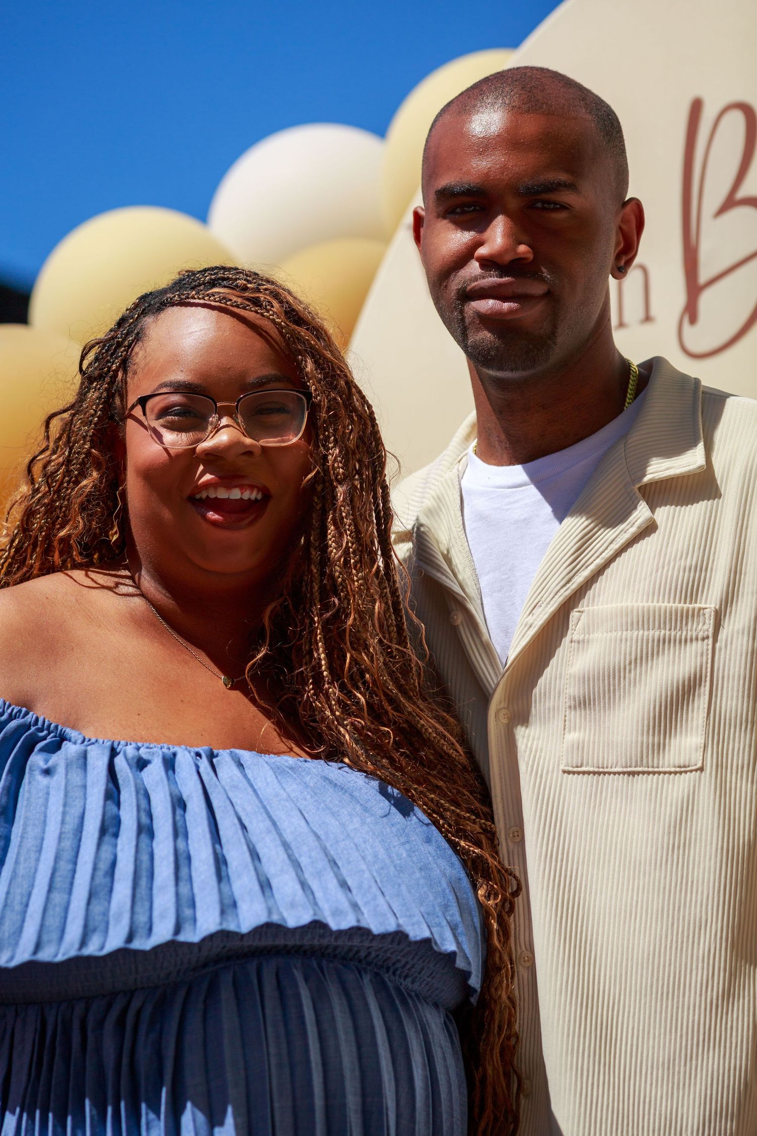A smiling woman with braids and glasses in a blue dress poses with a bald man in beige at an outdoor event.