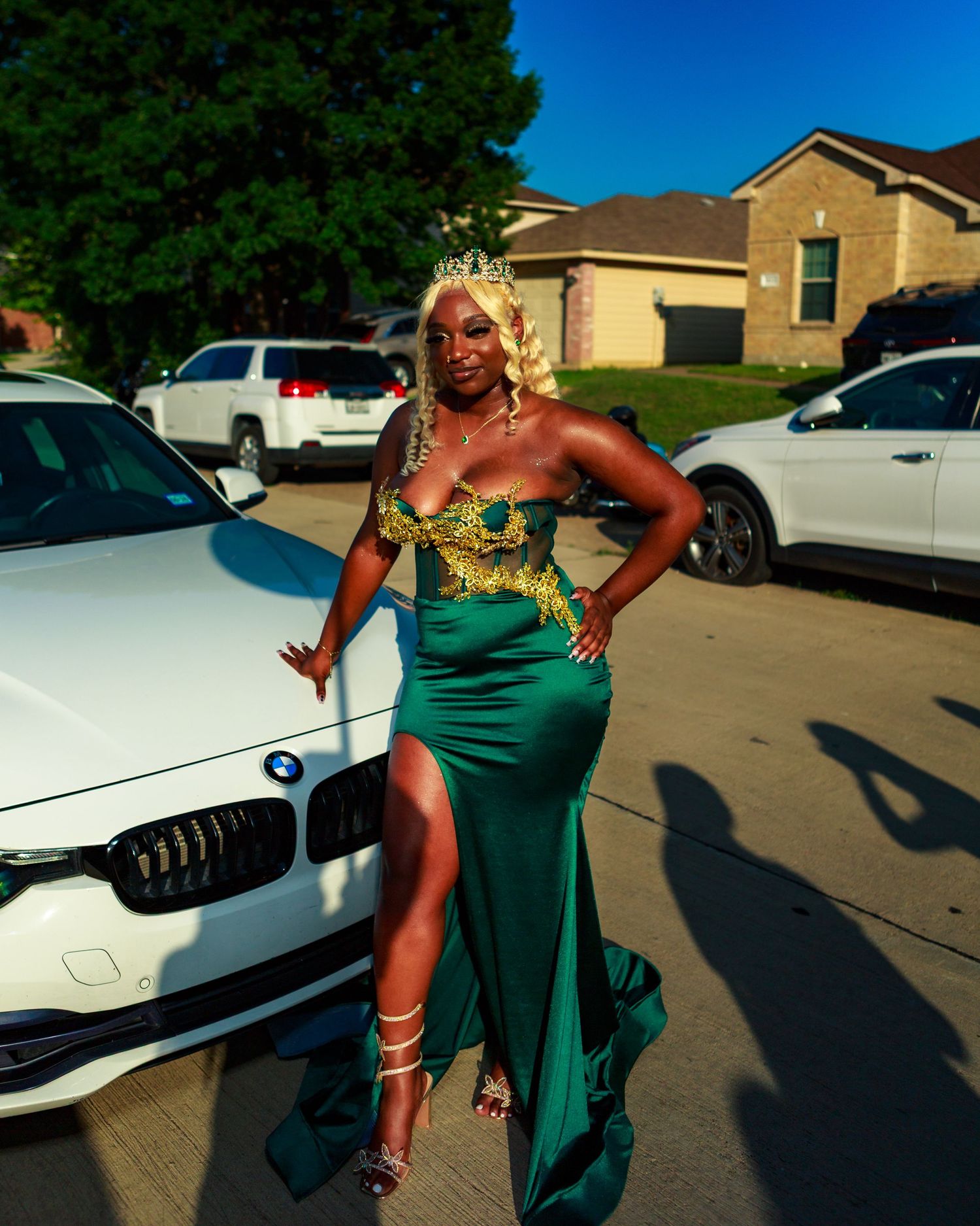 Woman in elegant green and gold gown with crown poses beside white BMW in suburban driveway.