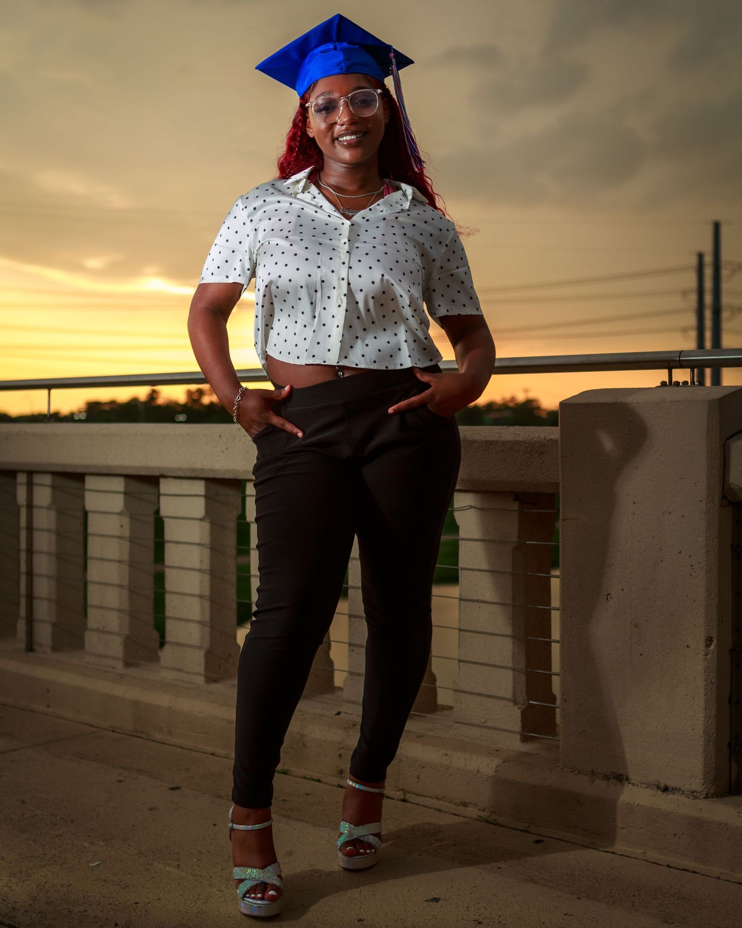 Graduate posing in blue cap, white shirt, and black pants at sunset.