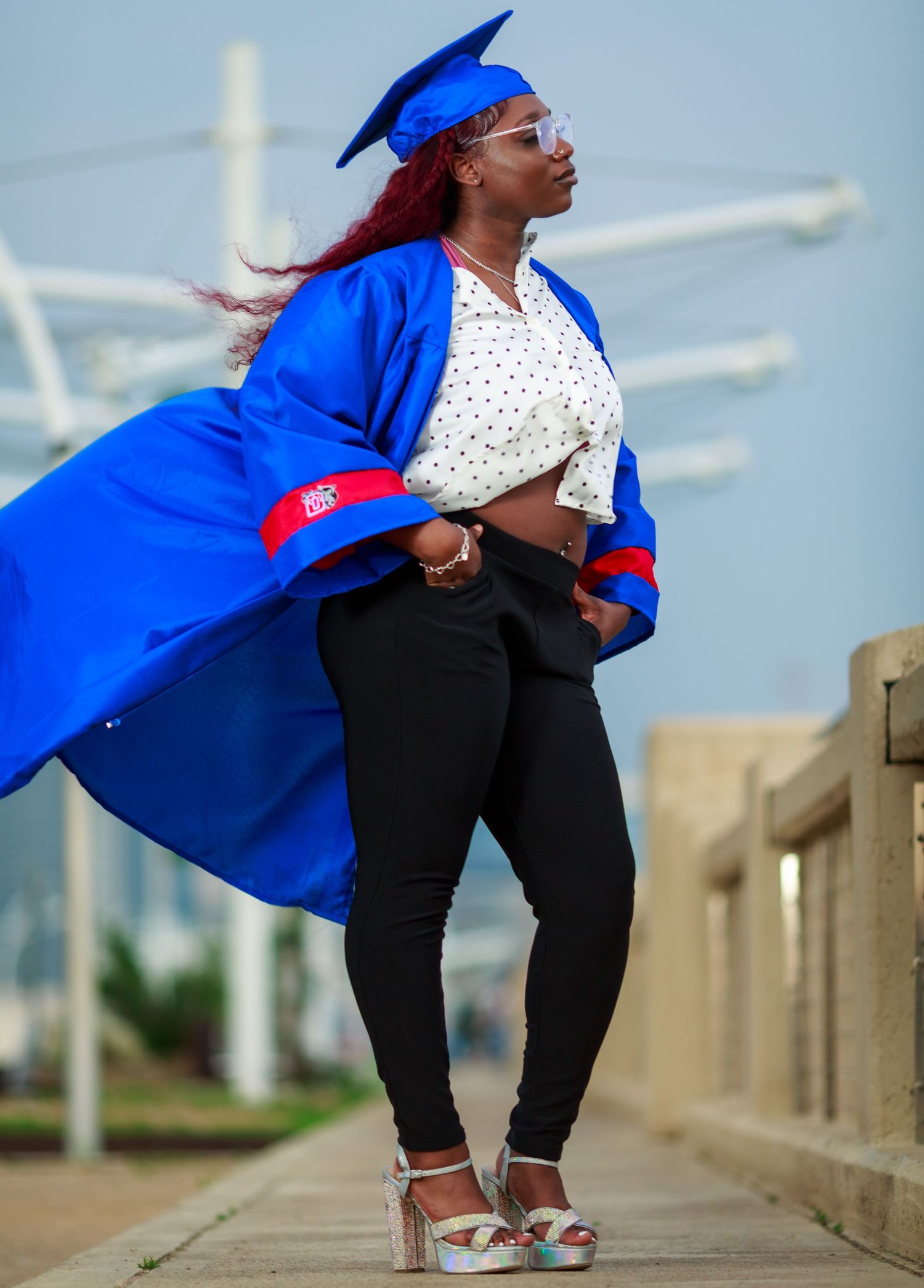 A woman in a blue graduation cap and gown poses confidently on a walkway, wearing black jeans and heels.