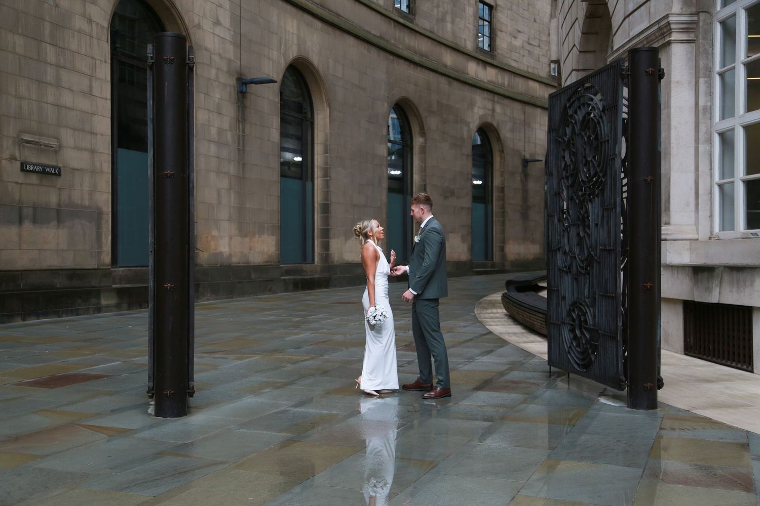 Bride and groom holding hands in a grand stone courtyard with ornate black gates on their wedding day. Ruby Salmon Photography