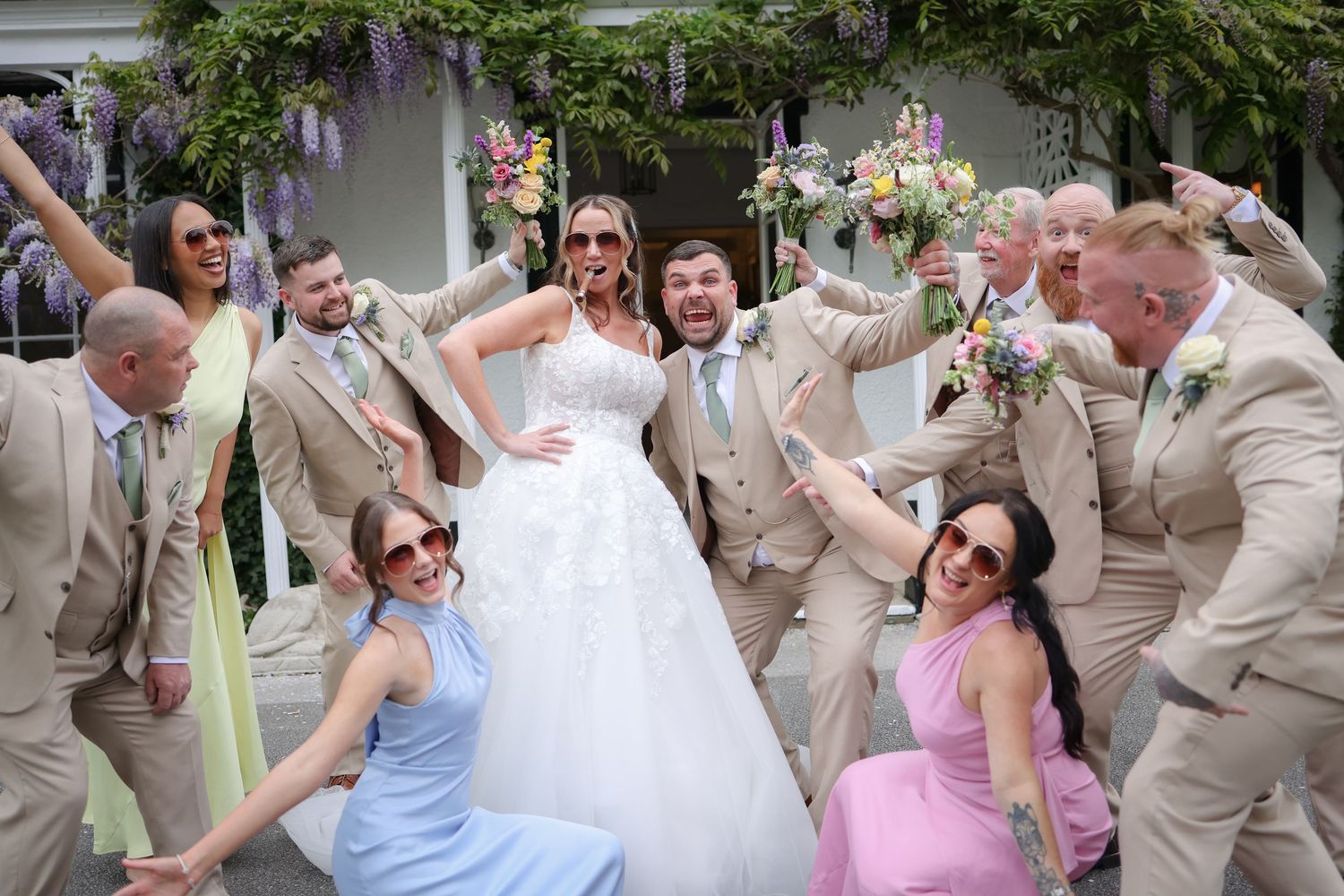Fun wedding party celebrating outdoors, bride in white gown surrounded by groomsmen in tan suits and bridesmaids in pastel dresses.