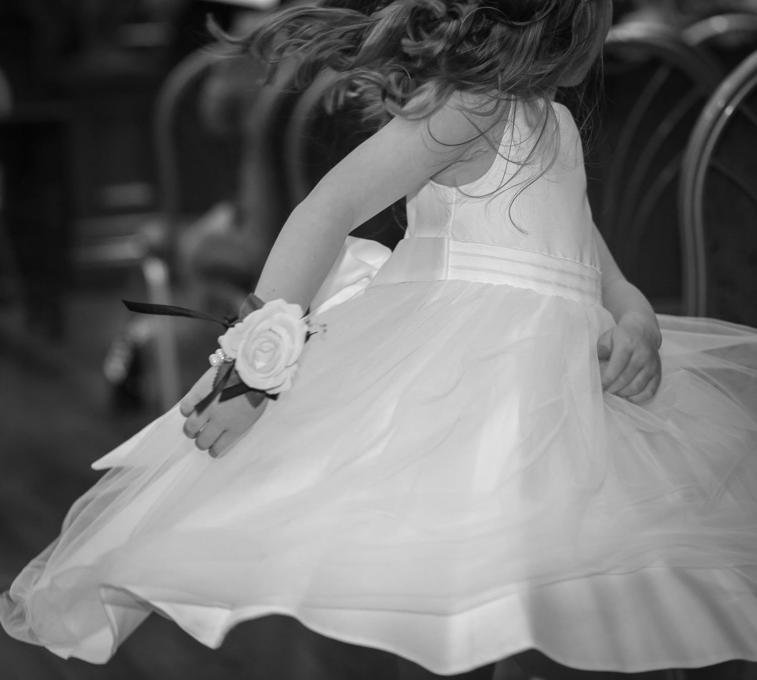 Black and white photo of a young flower girl in a white dress spinning gracefully, wearing a floral wrist corsage. Ruby Salmon Photography