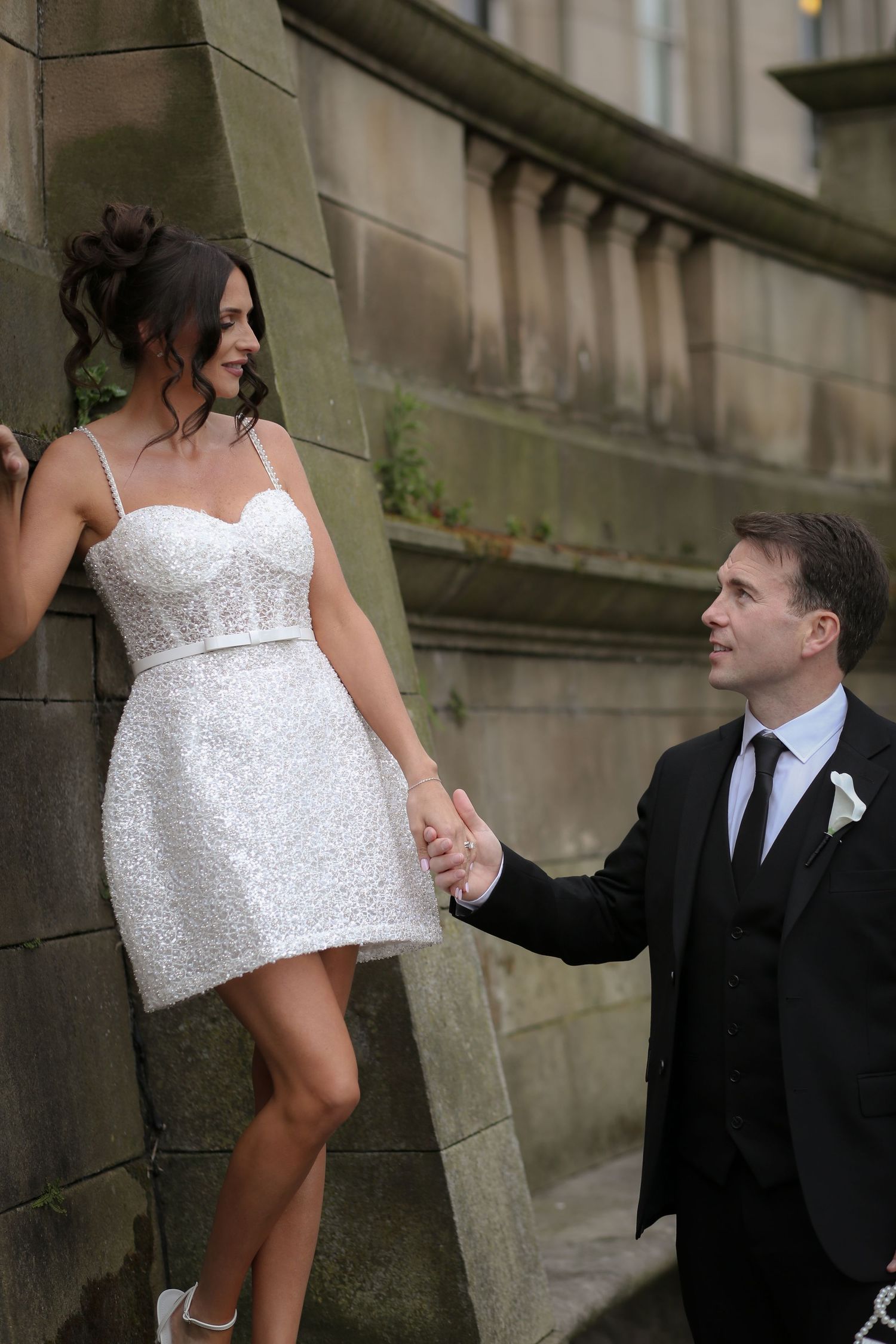 Bride in short white sequin dress holds hands with groom in black suit against stone balustrade. St Georges Hall, Liverpool