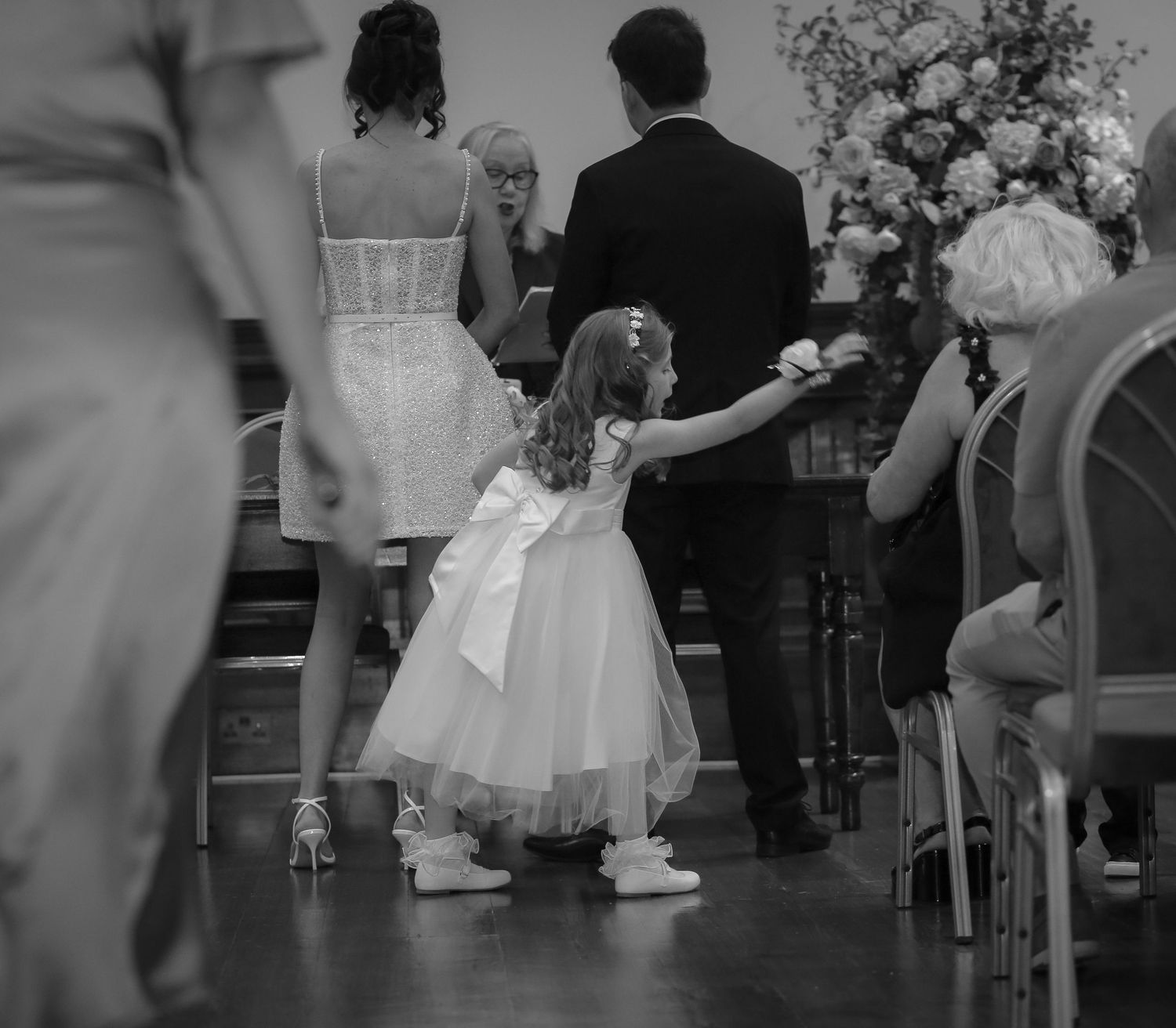 Black and white photo of a flower girl in a white dress reaching out during a wedding ceremony. St Georges Hall, Liverpool. Ruby Salmon