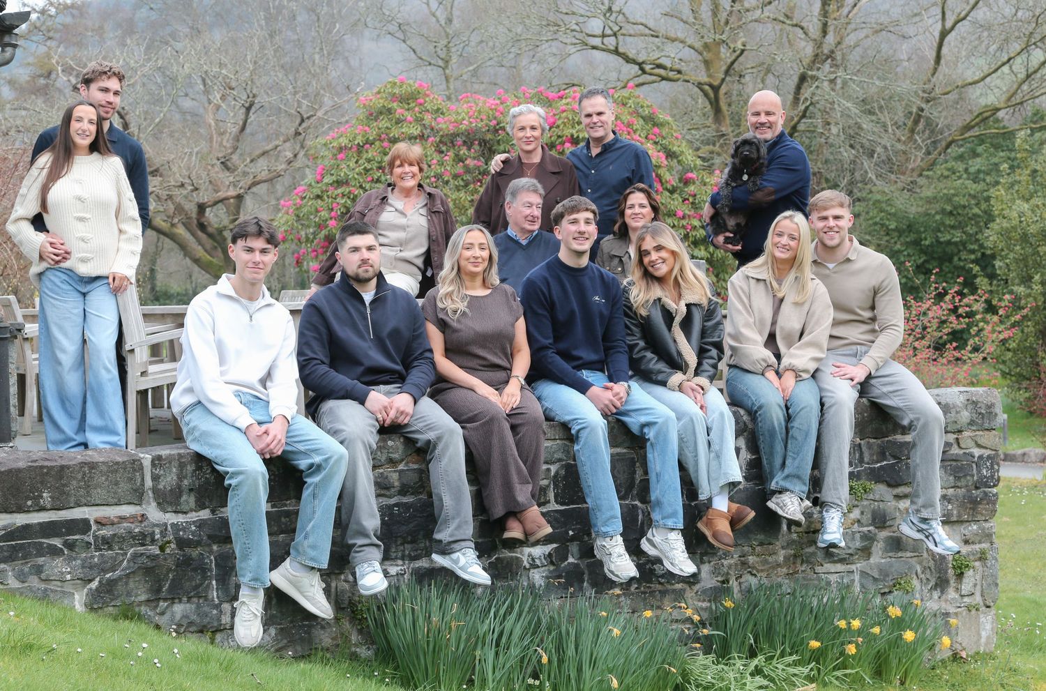 A large family group poses together on a stone wall in a lush garden setting with trees and daffodils in the background. Tan Llan, N. Wales