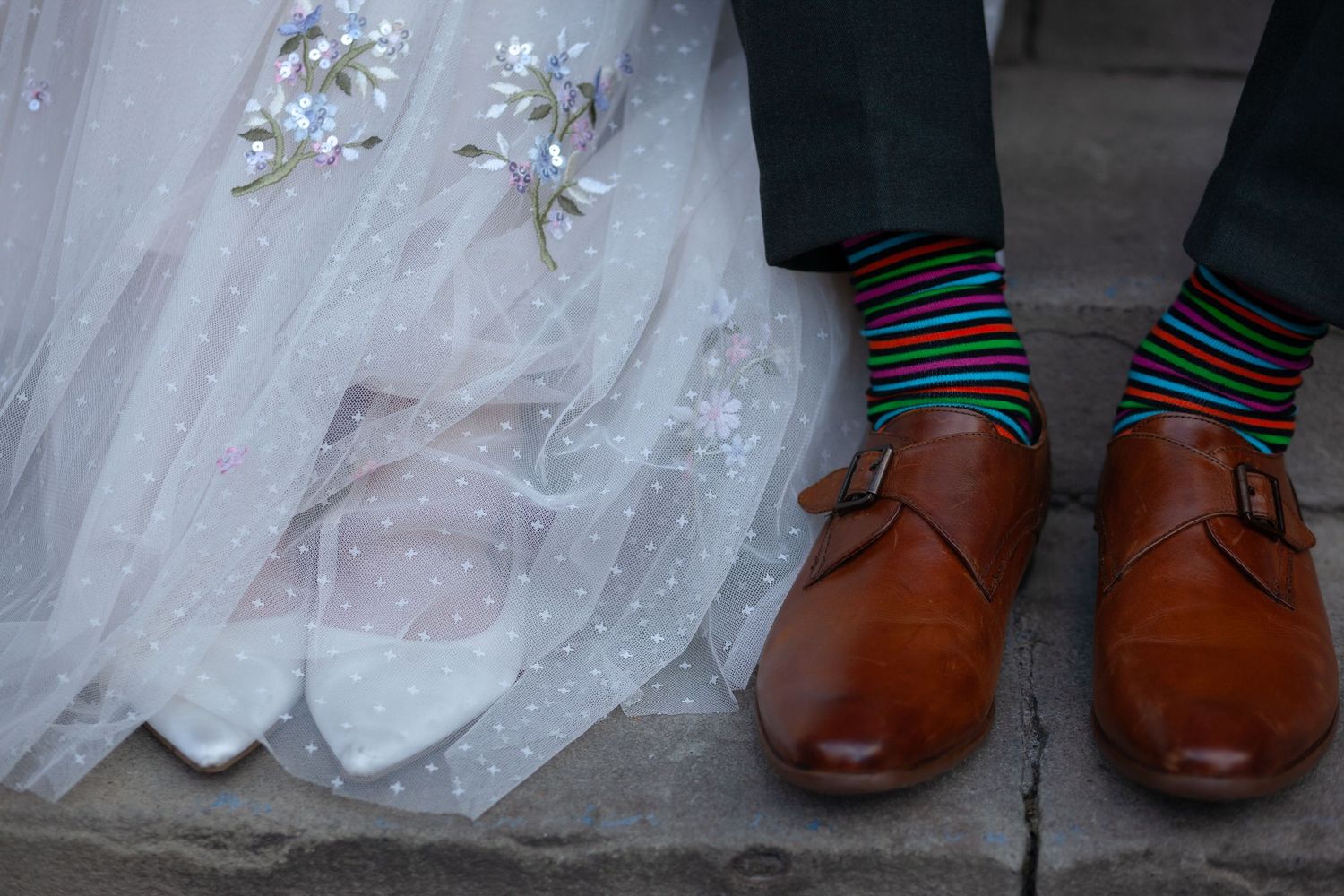 Close-up of bride's white heels and groom's brown monk strap shoes with colorful rainbow striped socks at wedding. Ruby Salmon Photography