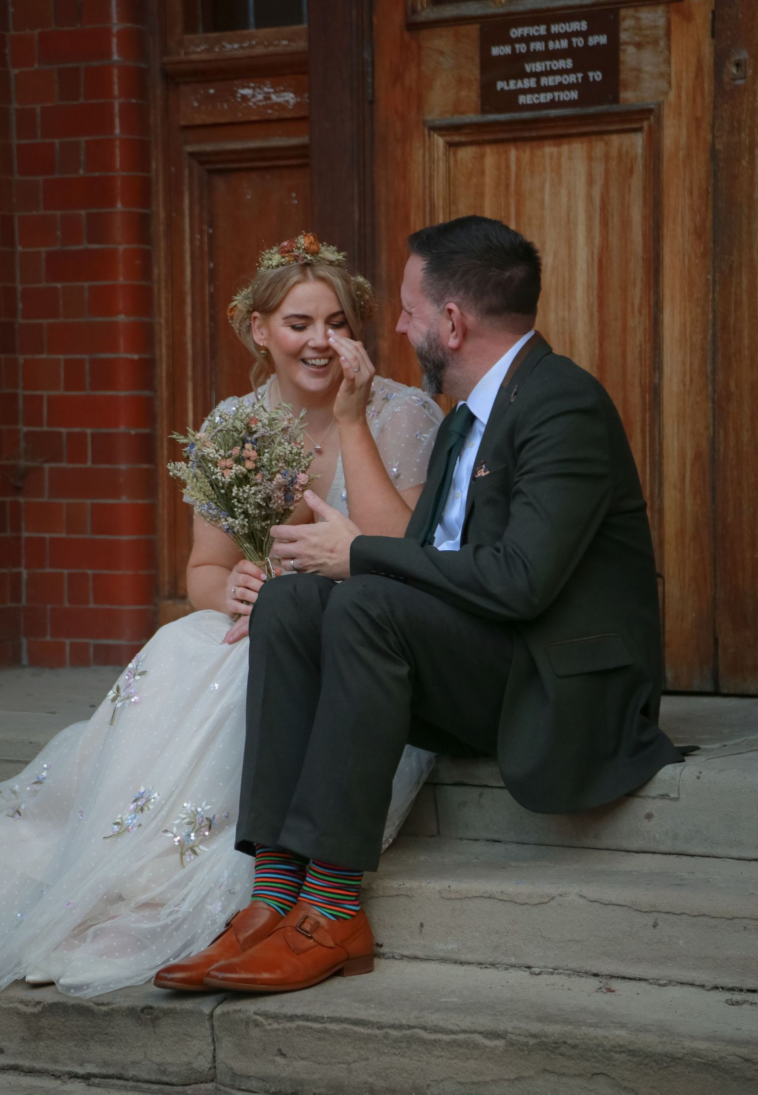 A laughing bride in a floral gown sits with a suited groom on steps, sharing a joyful wedding moment outdoors. Ruby Salmon Photography