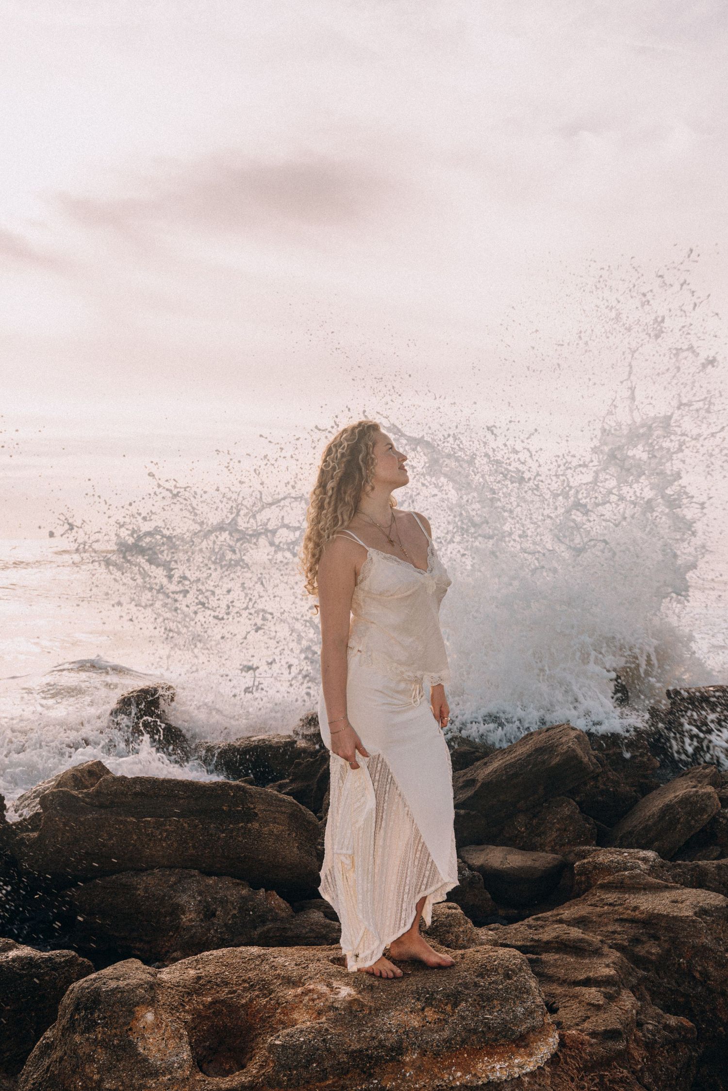Woman in white dress standing on rocks with crashing ocean waves behind her.