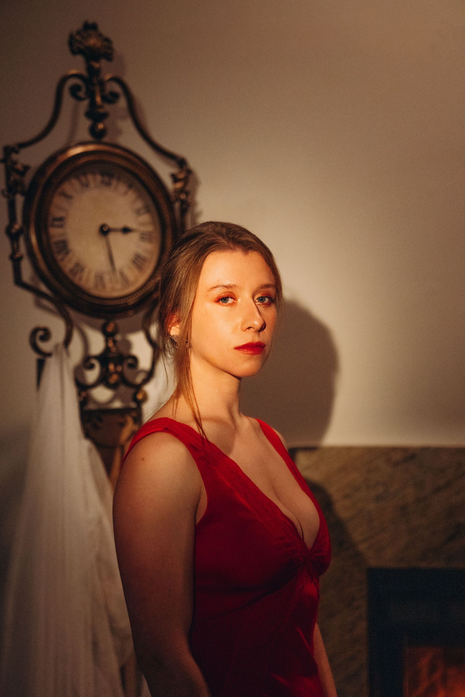 Elegant woman in red velvet dress posing near vintage gold clock and fireplace in warm light.