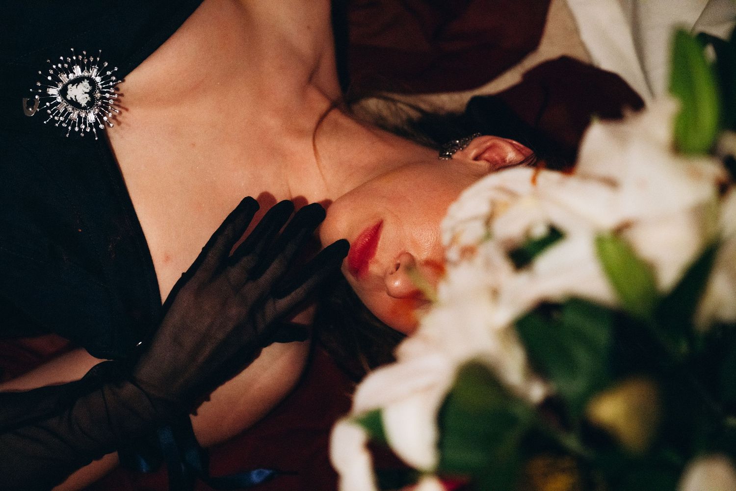 Woman in black gloves lying beside white flowers, wearing red lipstick in dramatic moody lighting.