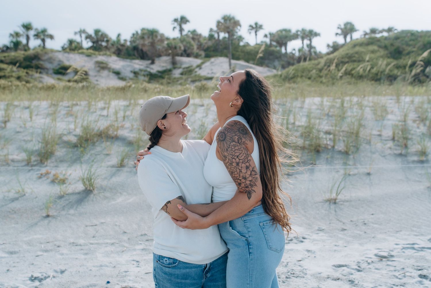 A couple embraces joyfully on a sandy beach with palm trees and dunes in the background.