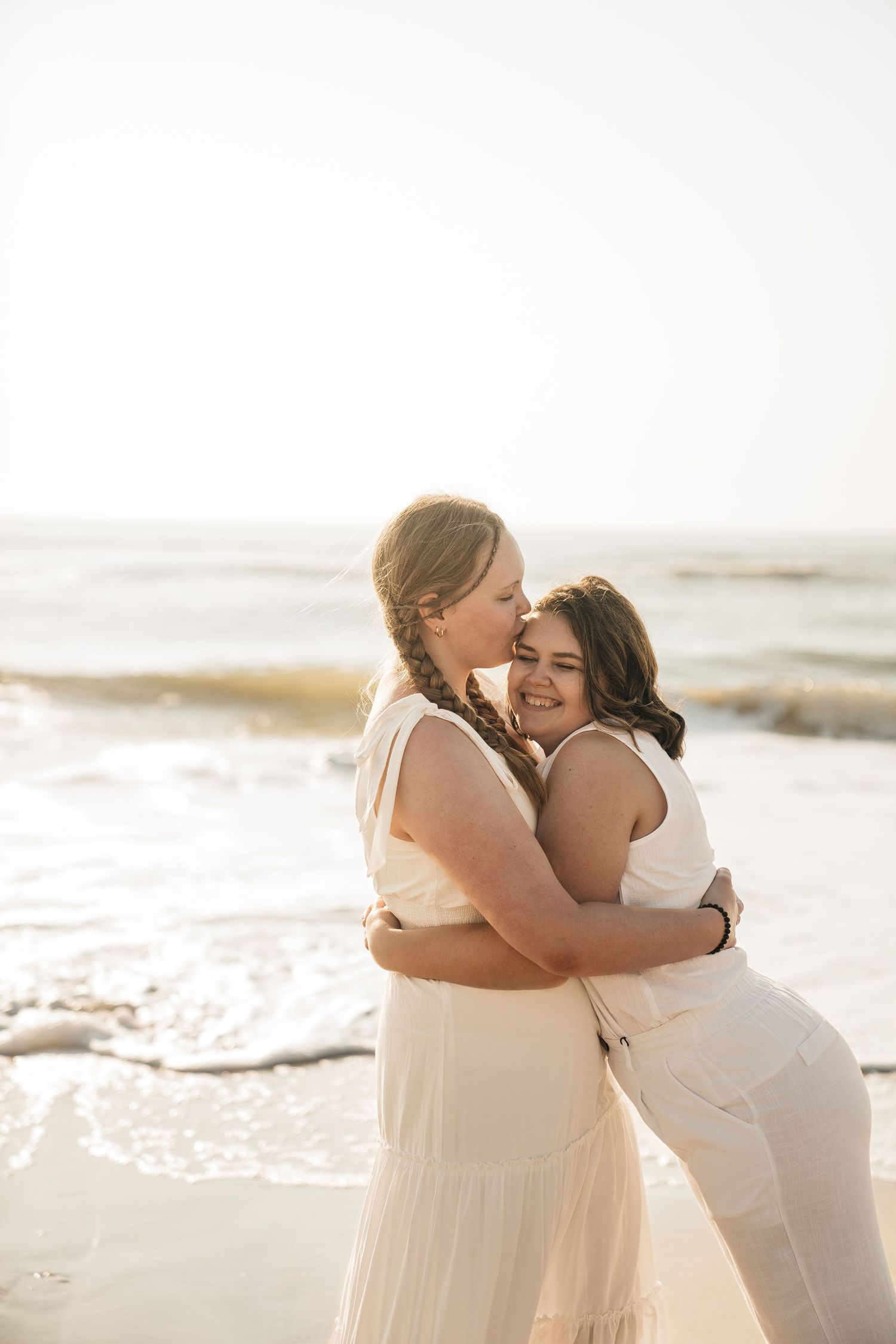 Two people in white dresses sharing a joyful embrace on the beach.