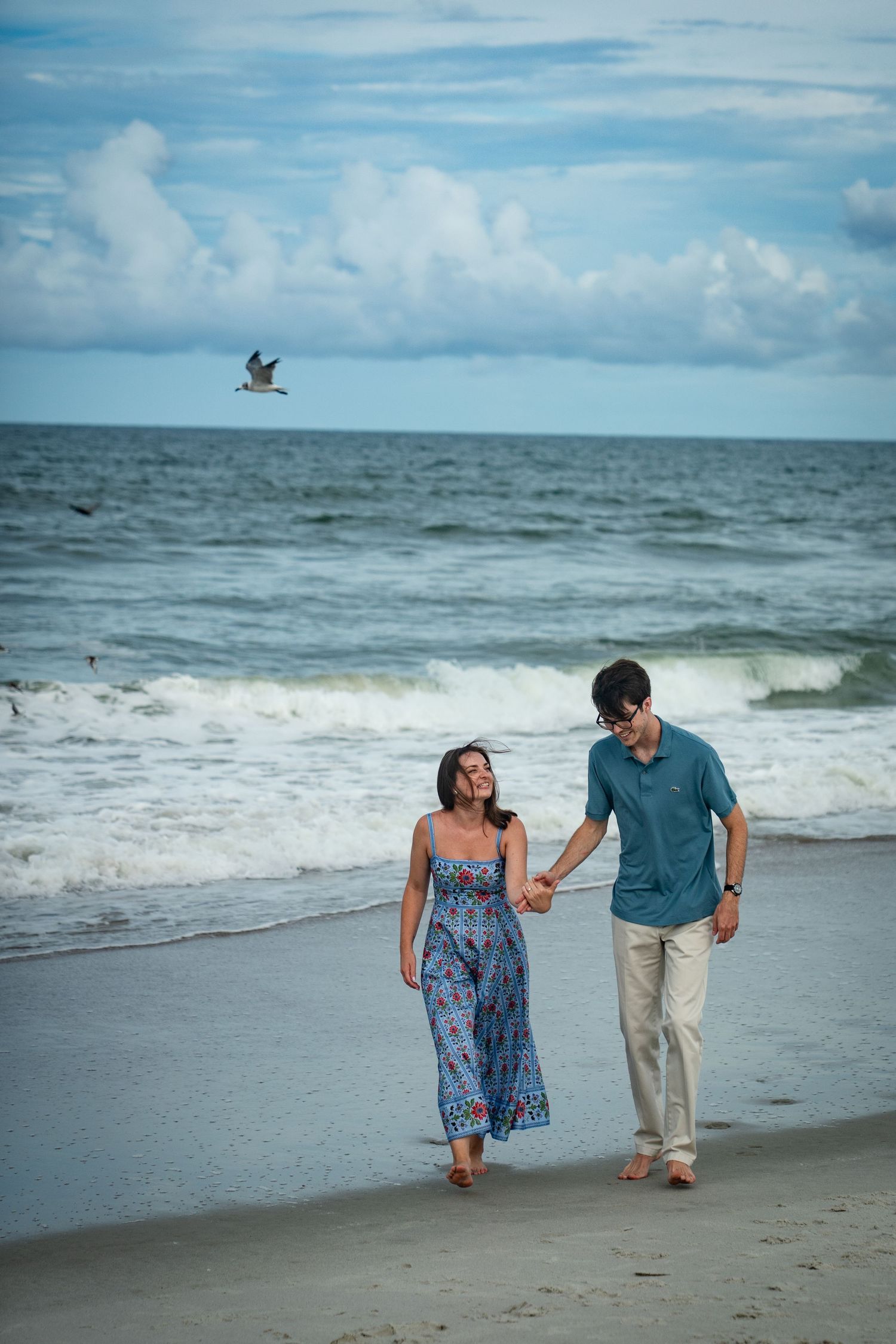 A couple walks hand in hand along a scenic beach with waves crashing and seagulls flying overhead.