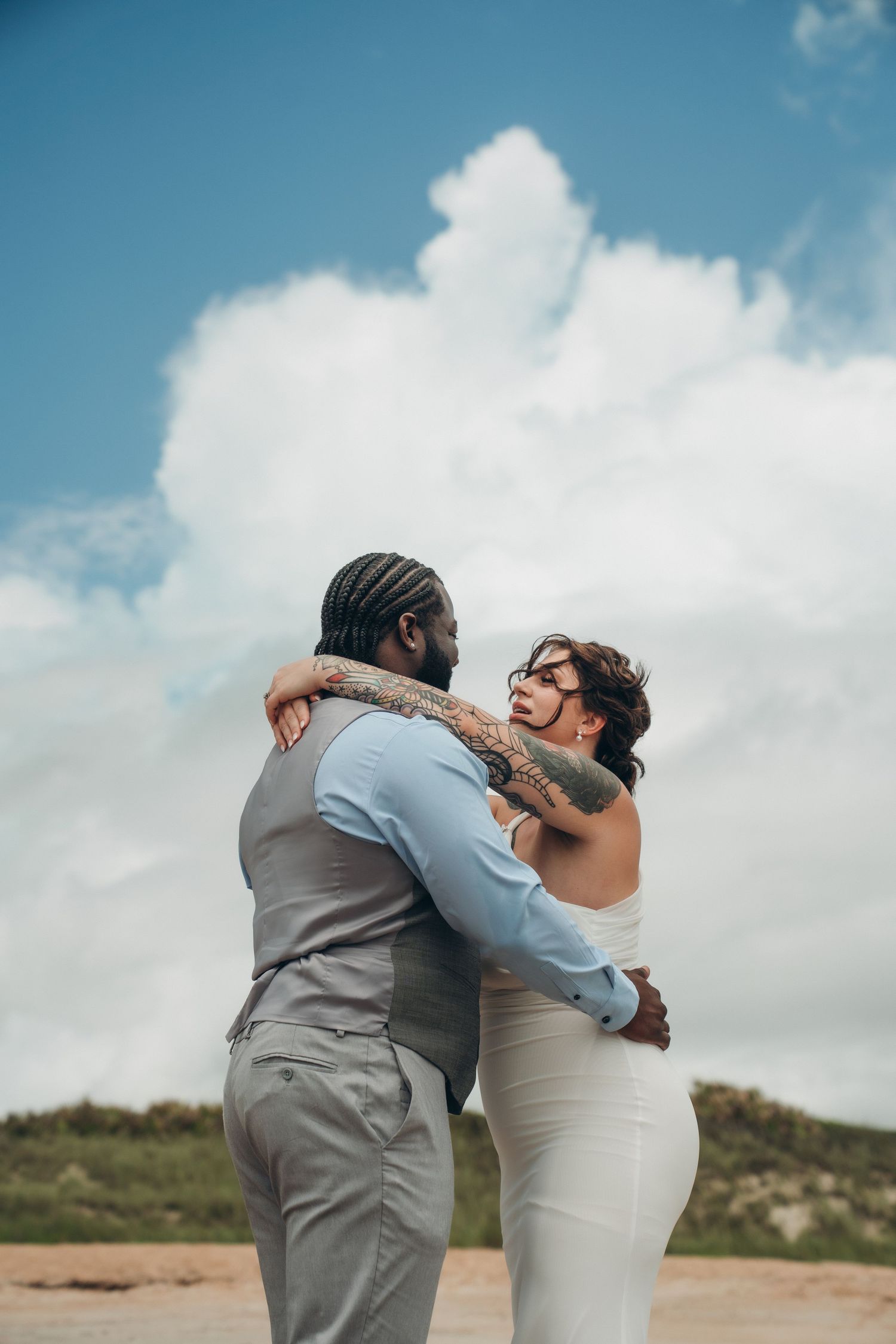 Interracial couple in wedding attire embracing outdoors under a blue sky with clouds, groom in gray suit, bride in white dress.
