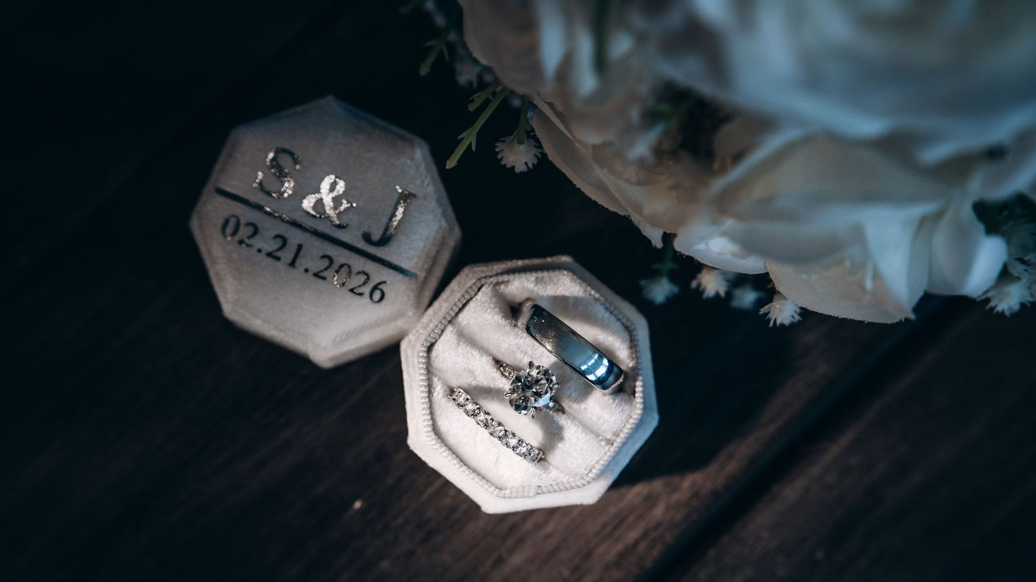 Elegant wedding ring box engraved with E&J and date 02.21.2026, displaying diamond engagement and wedding bands near white flowers.