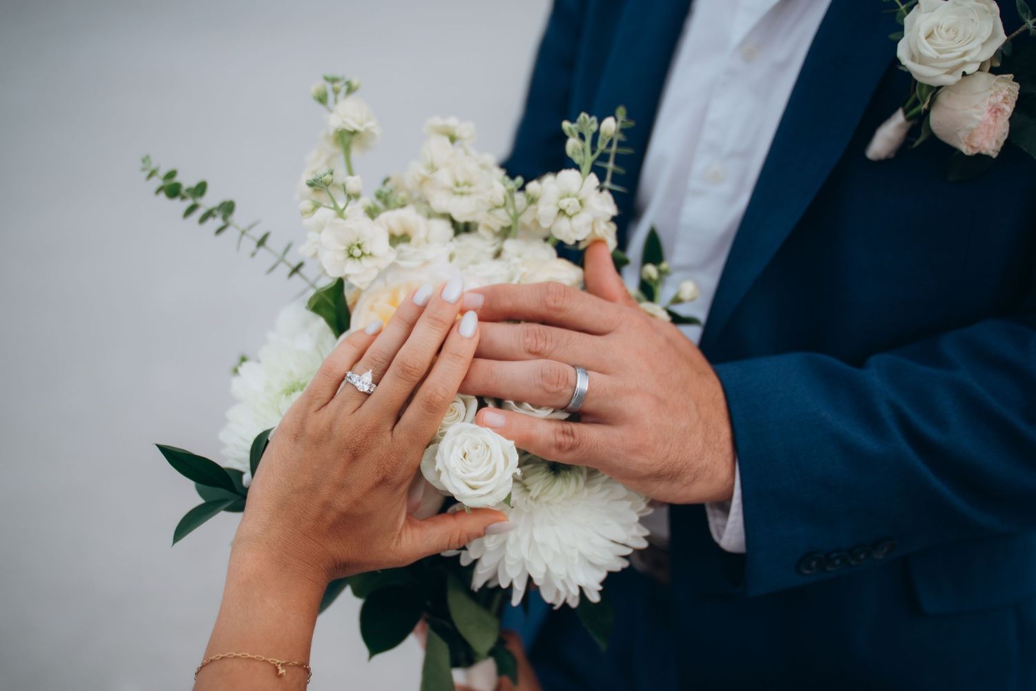 Bride and groom hands with wedding rings resting on white floral bouquet, groom in navy blue suit.